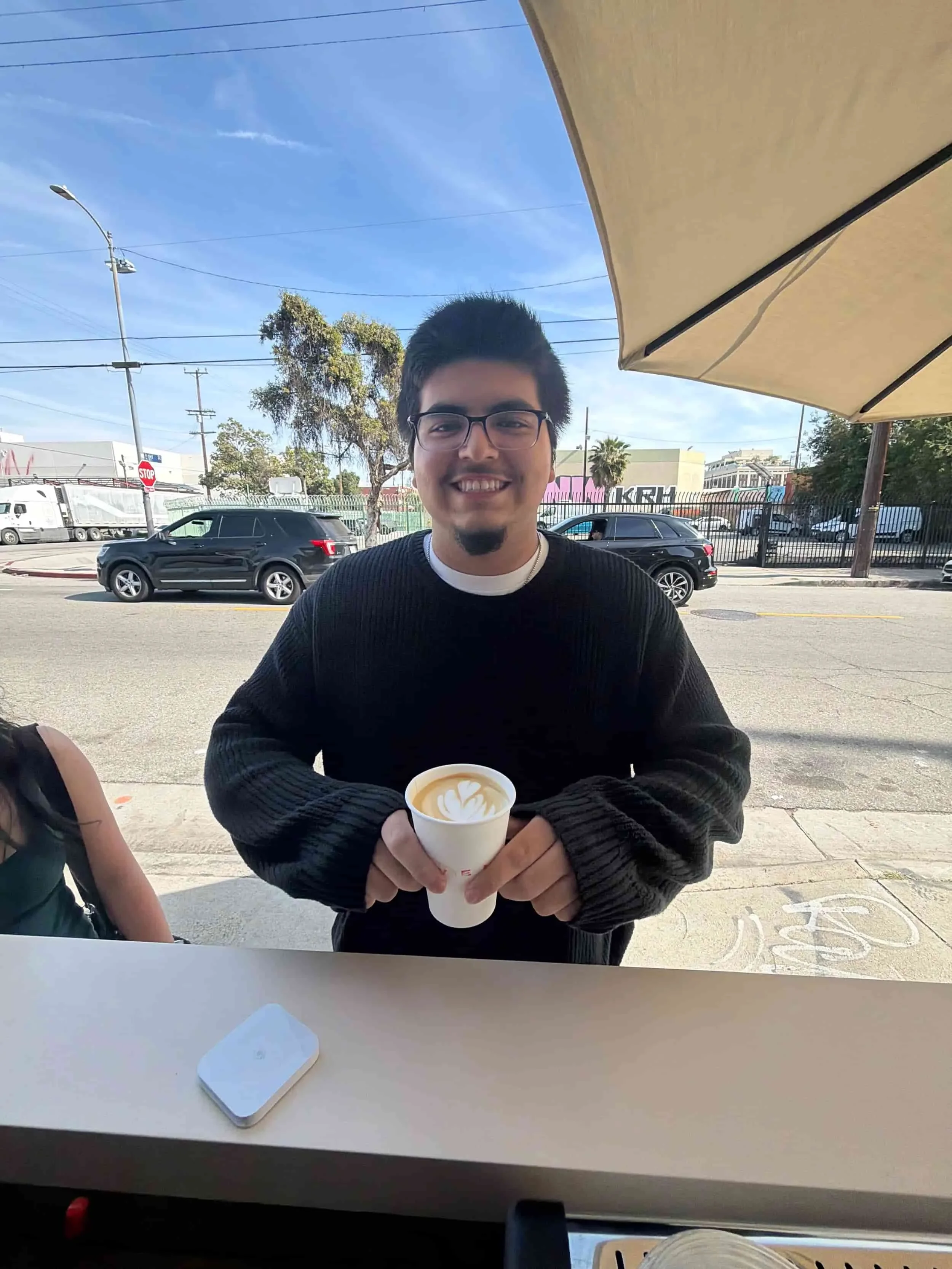 A man with black hair, glasses, and a goatee sitting outside at a table holding a cup of coffee with latte art. He is smiling and wearing a black sweater and a white shirt underneath. In the background, there are cars driving on the street, trees, po