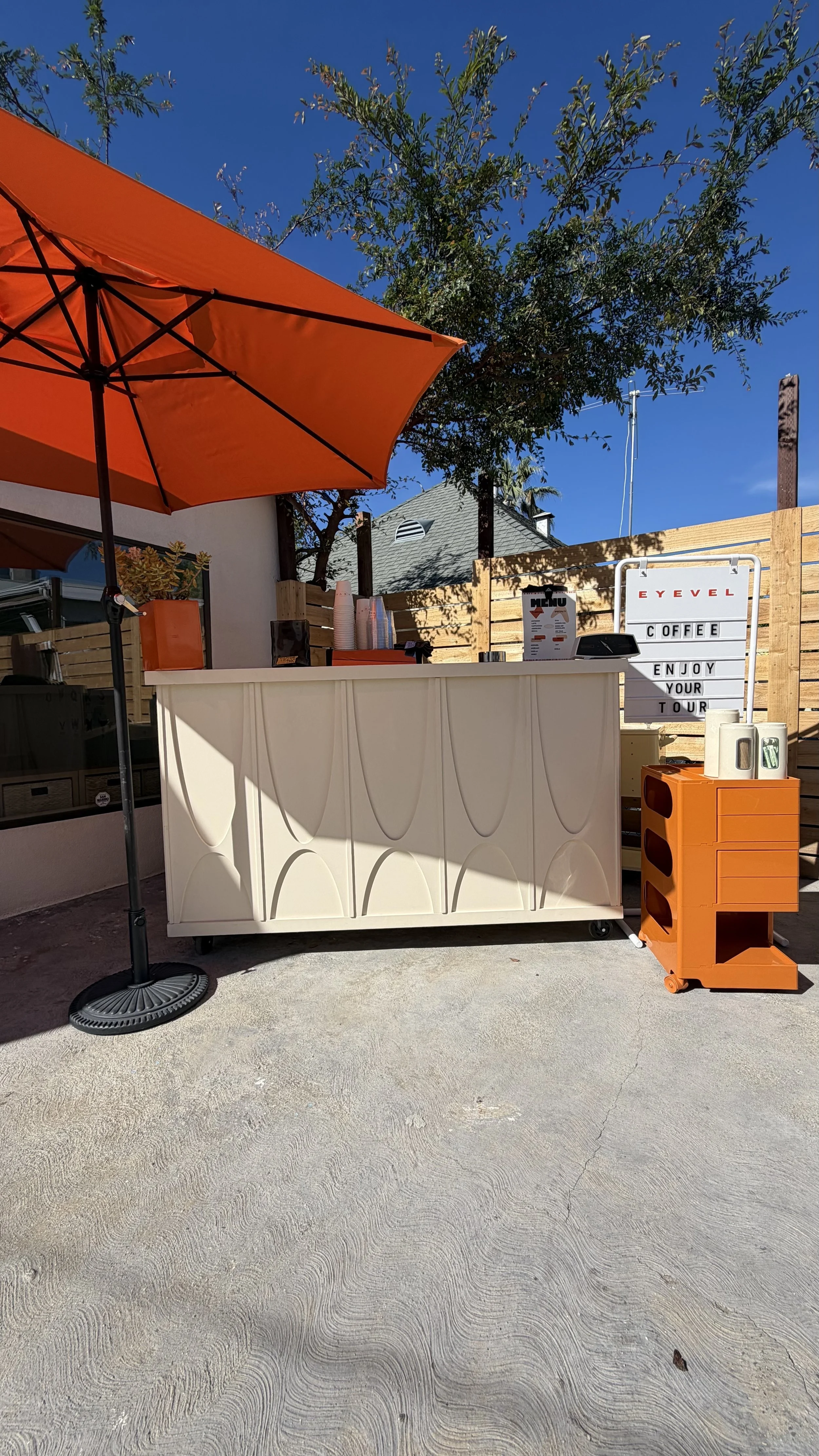 Outdoor coffee stand with orange umbrella, white counter, and wooden fence in the background under clear blue sky.
