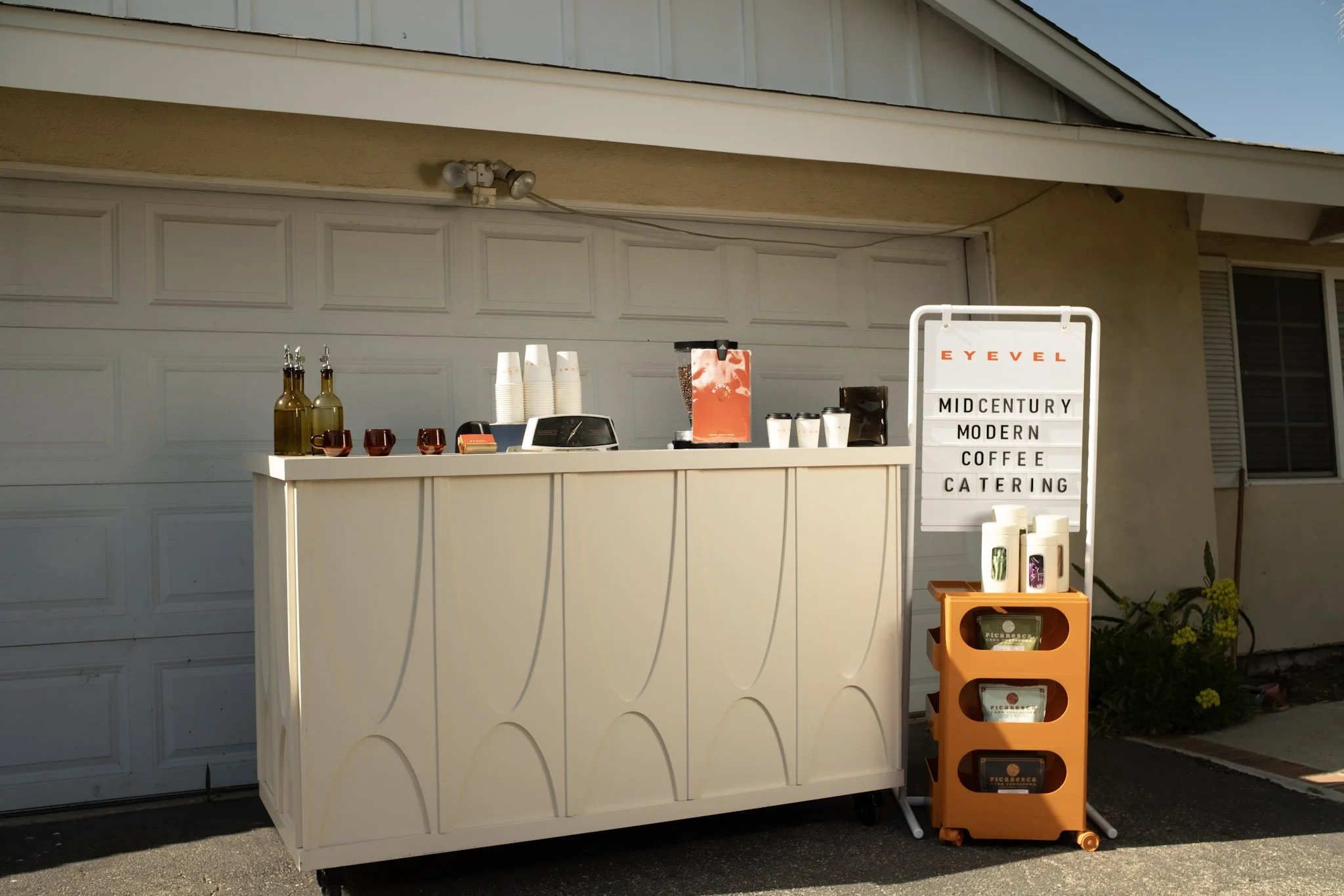 Outdoor coffee stand with a white counter, coffee cups, bottles, and a sign that reads 'Midcentury Modern Coffee Catering' at EYEVEL company, situated in front of a house garage.