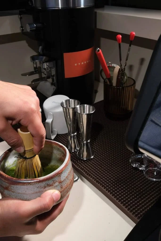 Person mixing matcha tea with a bamboo whisk in a ceramic bowl on a coffee station countertop.