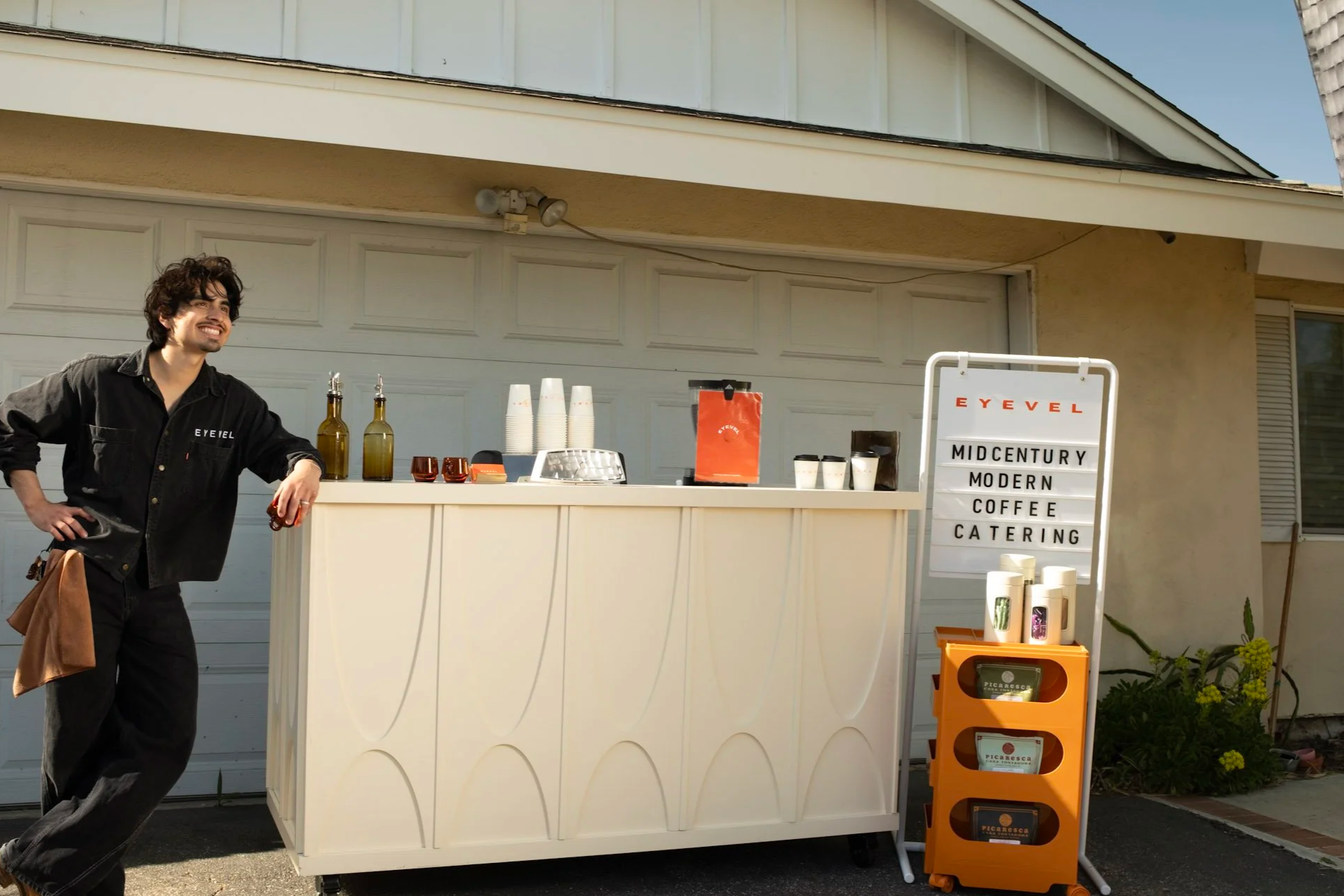 A smiling man with dark curly hair standing near a coffee stand outside a building, with a sign that reads 'Midcentury Modern Coffee Catering.'