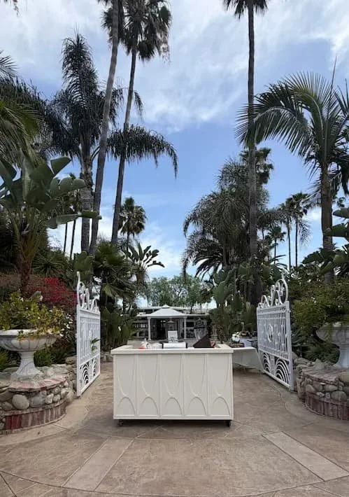 A backyard with tall palm trees, lush green plants, and a white garden gate. A white outdoor island with a sink and a small table is in the center, with a partly cloudy sky overhead.