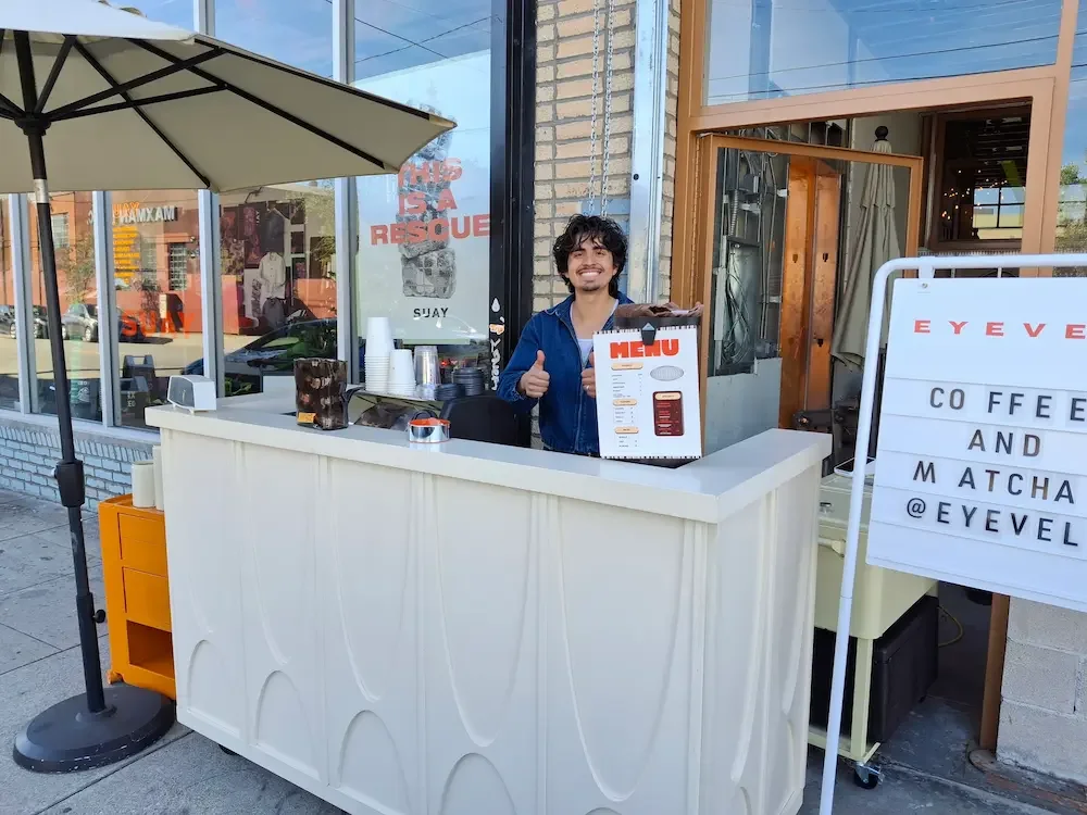 A person stands behind an outdoor coffee stand in Pasadena, holding a menu and giving a thumbs up—perfect for coffee catering events.