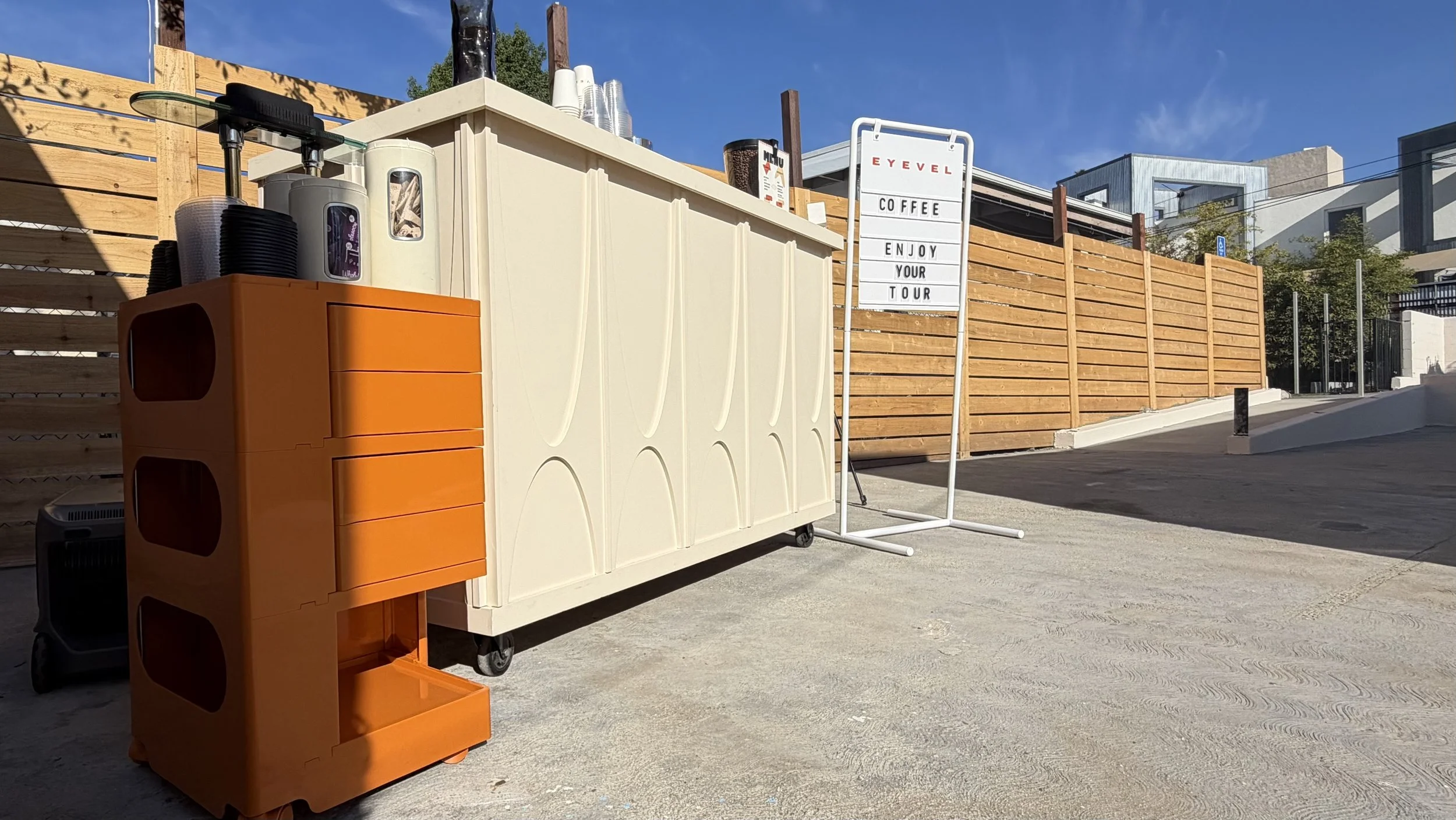 Outdoor scene with a coffee stand, white signboard saying 'EYEVEL COFFEE ENJOY YOUR TOUR,' and wooden fence in the background under clear blue sky.