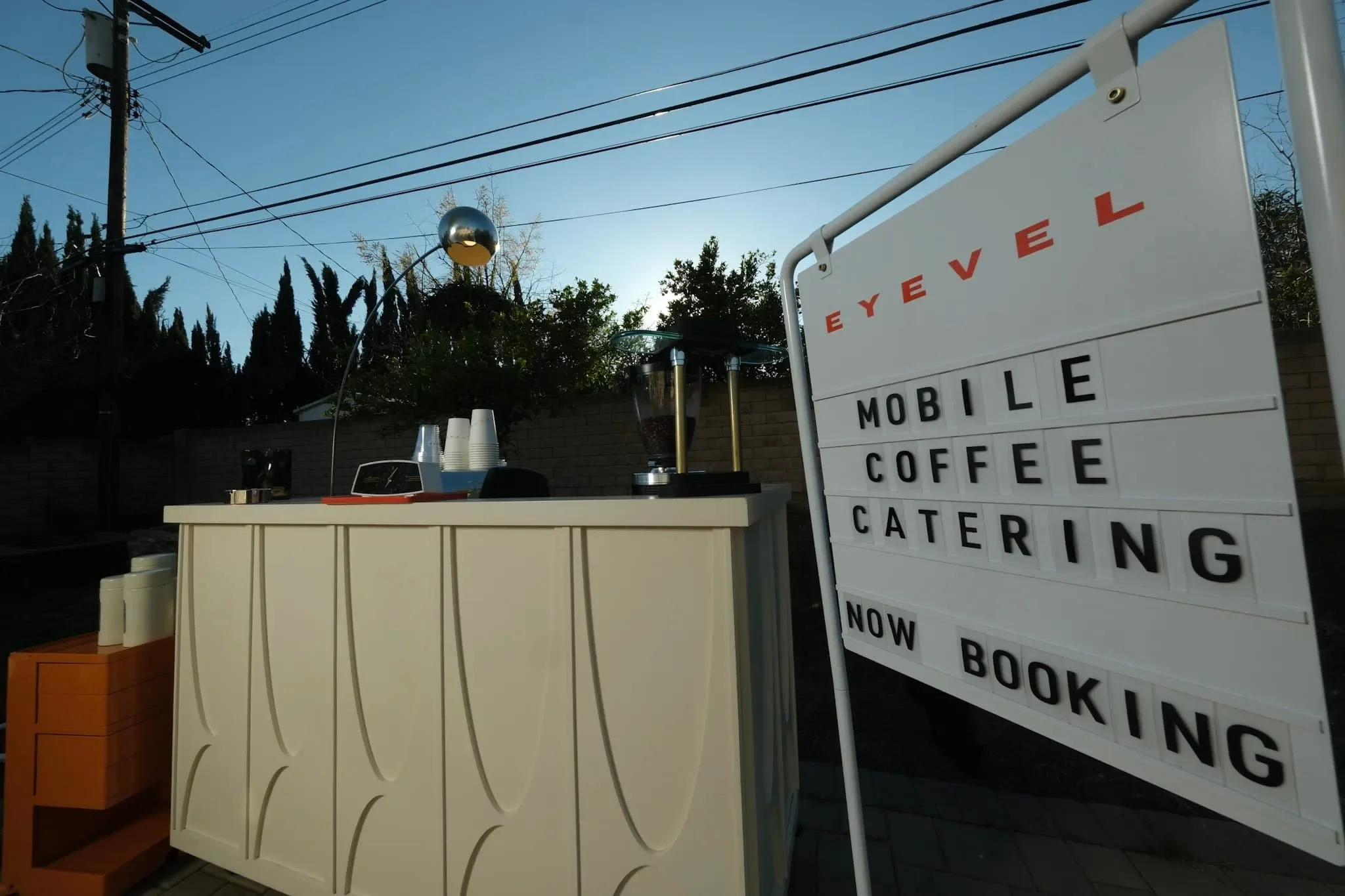 An outdoor coffee stand with a white counter, a coffee grinder, and cups. A large sign reads, 'EVE L,' 'MOBILE COFFEE,' 'CATERING,' and 'NOW BOOKING.' Power lines and trees are visible against a clear sky.