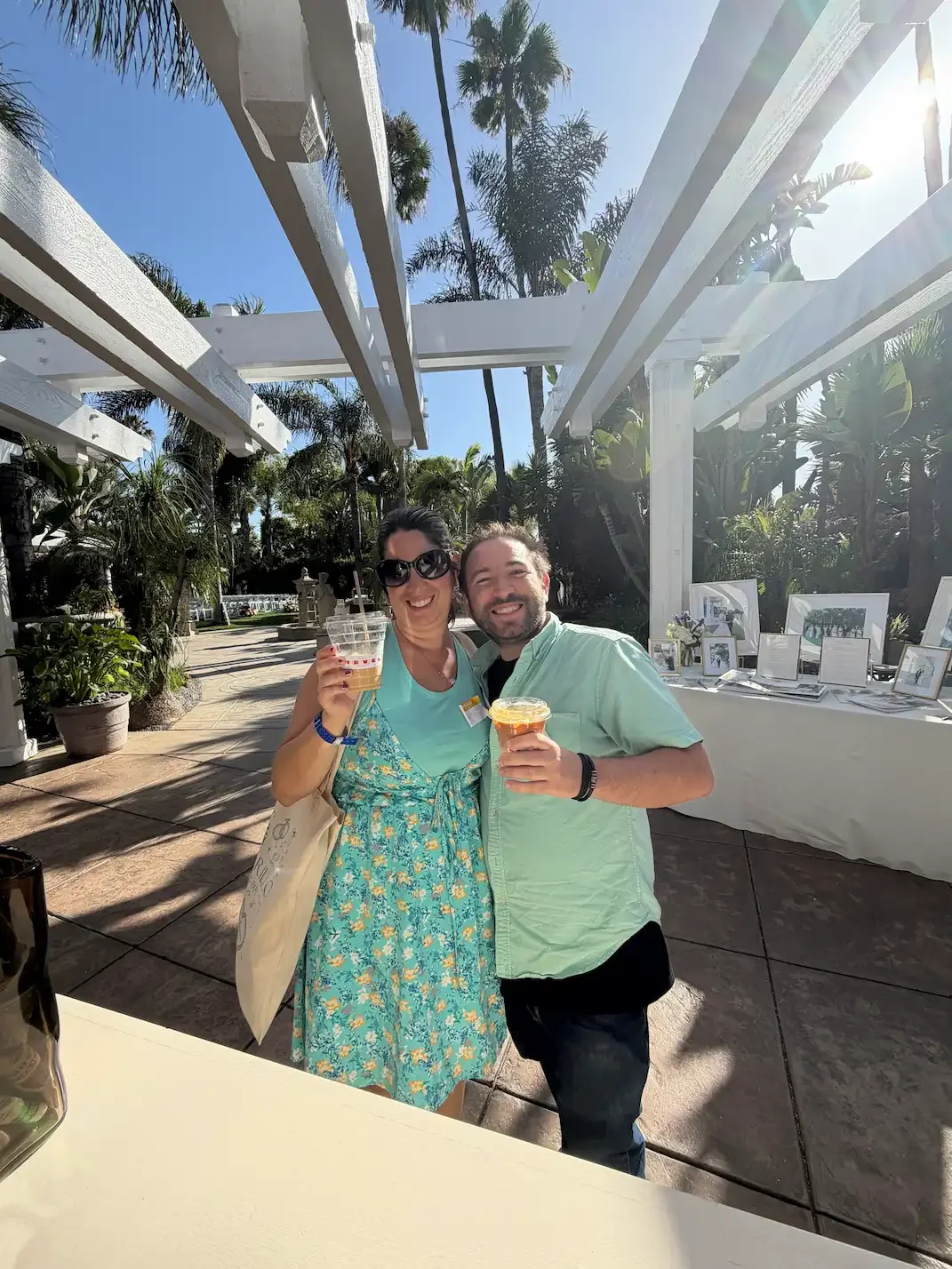 Two people smiling at an outdoor event, holding drinks, with a table displaying photographs and framed pictures in the background, under a white pergola with palm trees and bright sunlight.