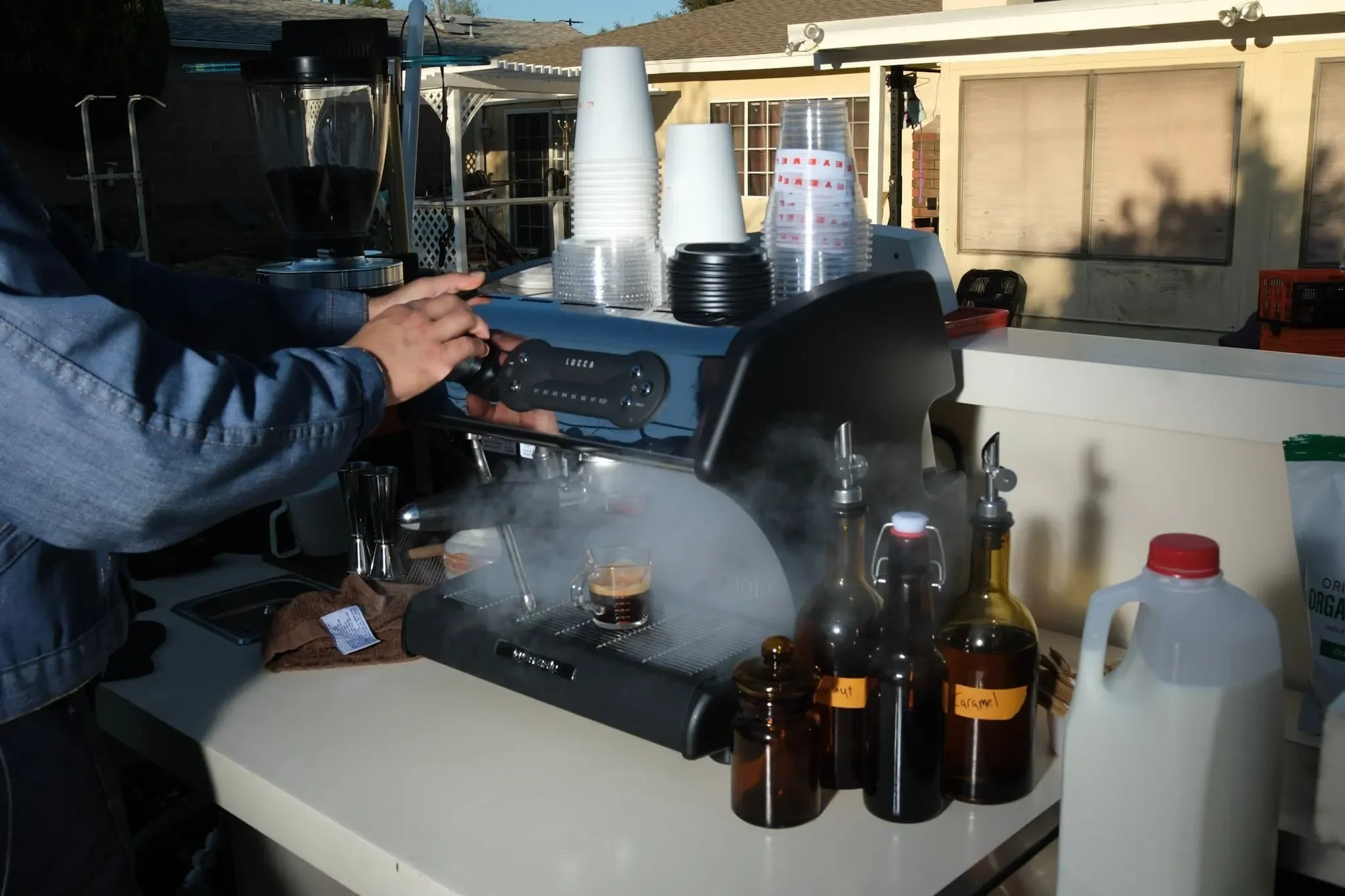 A person making coffee using a pour-over coffee maker on a counter outdoors. There are stacks of paper cups, coffee bottles, and other supplies on the counter. The background shows a house with a window and a sliding door.