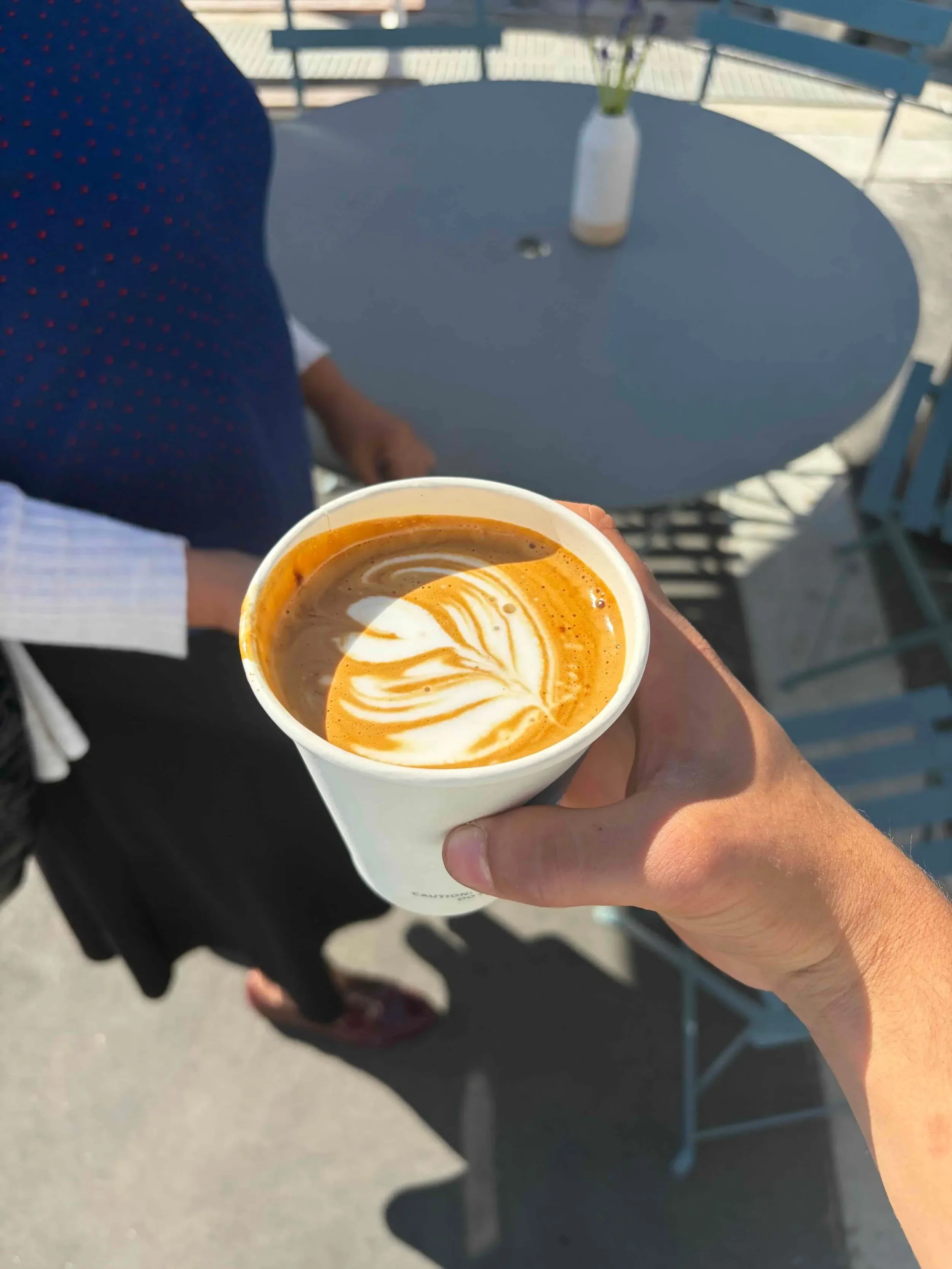 A person is holding a cup of coffee with latte art, outside in daylight, with a round table and vase with flowers in the background.