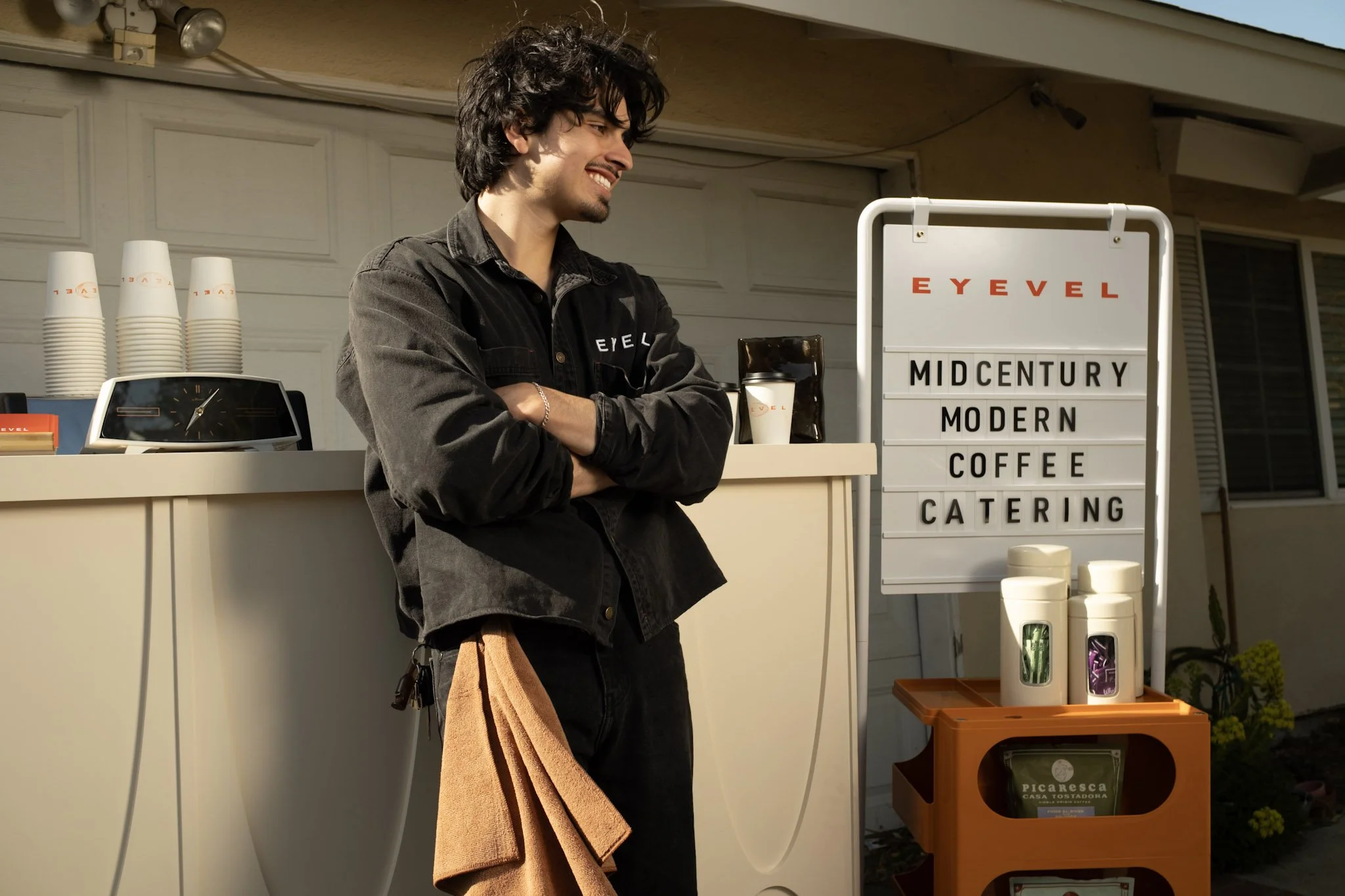 Young man standing outside a coffee stand with a sign reading 'Midcentury Modern Coffee Catering' in front of a garage, with disposable cups and a coffee bag visible.