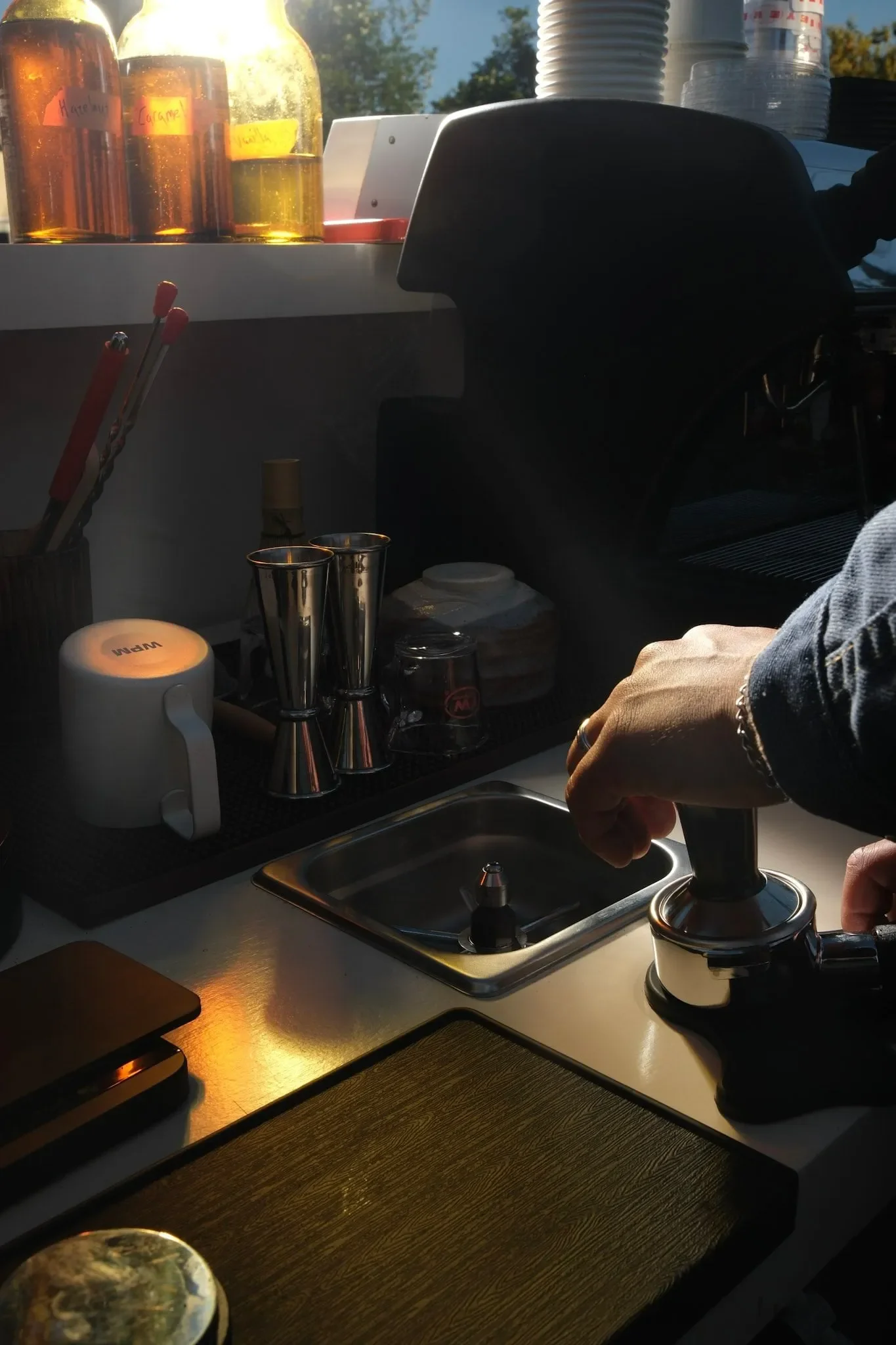 Close-up of a coffee shop counter with various bottles, a cup, mixing tools, and a person operating a coffee machine or grinder in the background. The scene is lit with warm sunlight.