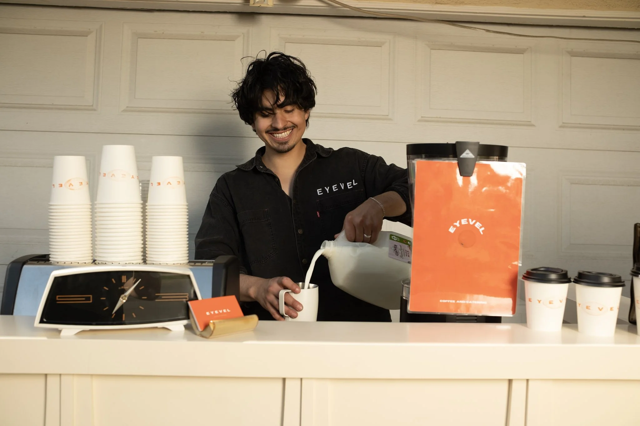 A barista in a black shirt with 'EYEVEL' logo is pouring milk into a mug at a coffee shop, with cups, coffee cups, and a coffee machine on the counter.