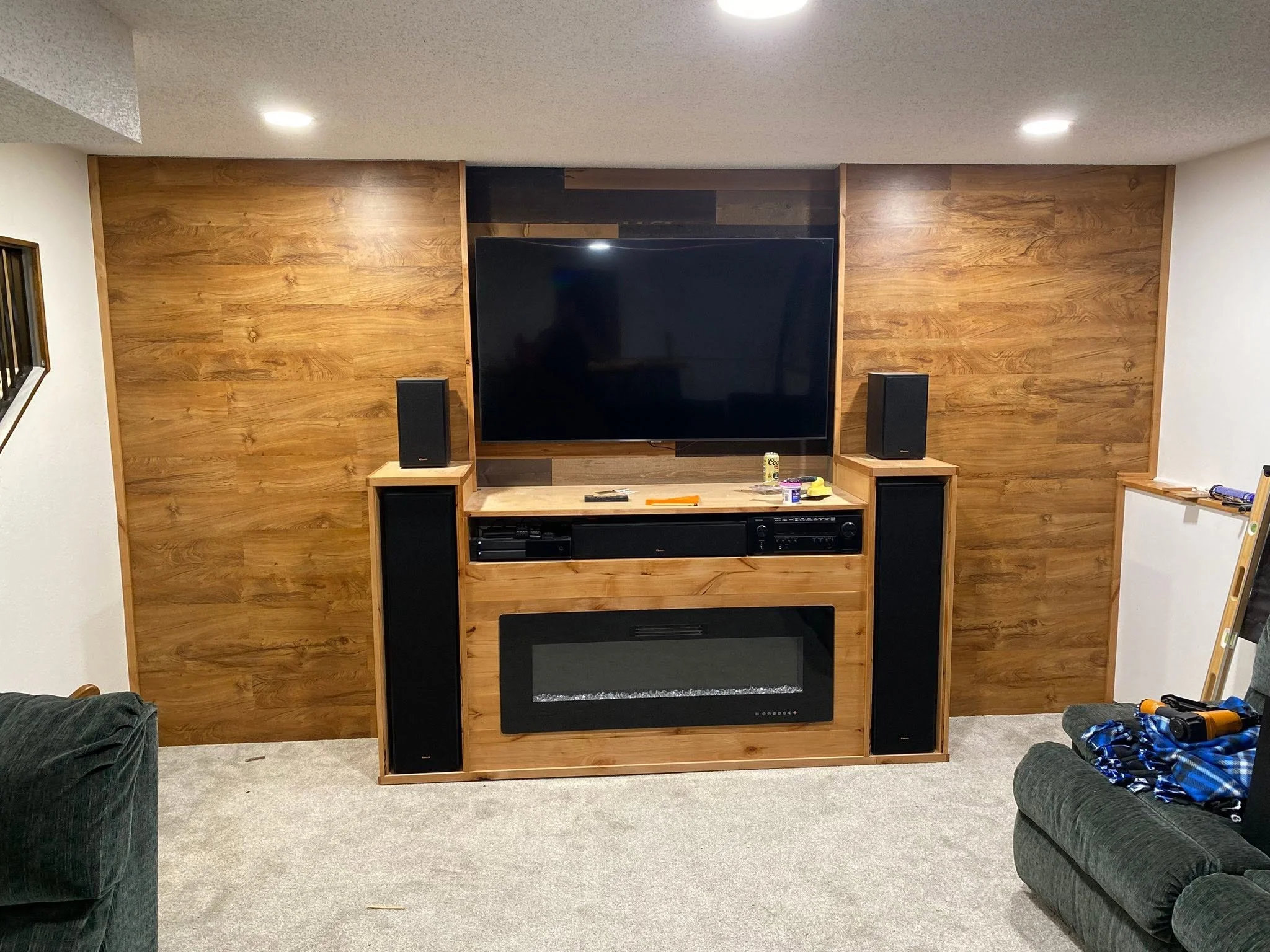 Living room with a large wooden wall unit containing a TV, speakers, and electronic devices, with furniture and clutter in the foreground.