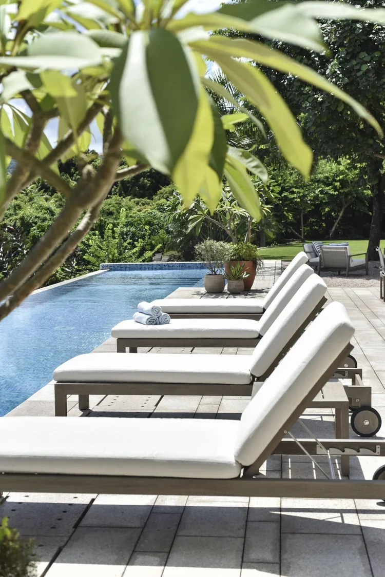 Serene poolside sanctuary featuring modern white chaise lounges with plush cushions beside a pristine blue pool, framed by lush tropical foliage and verdant gardens