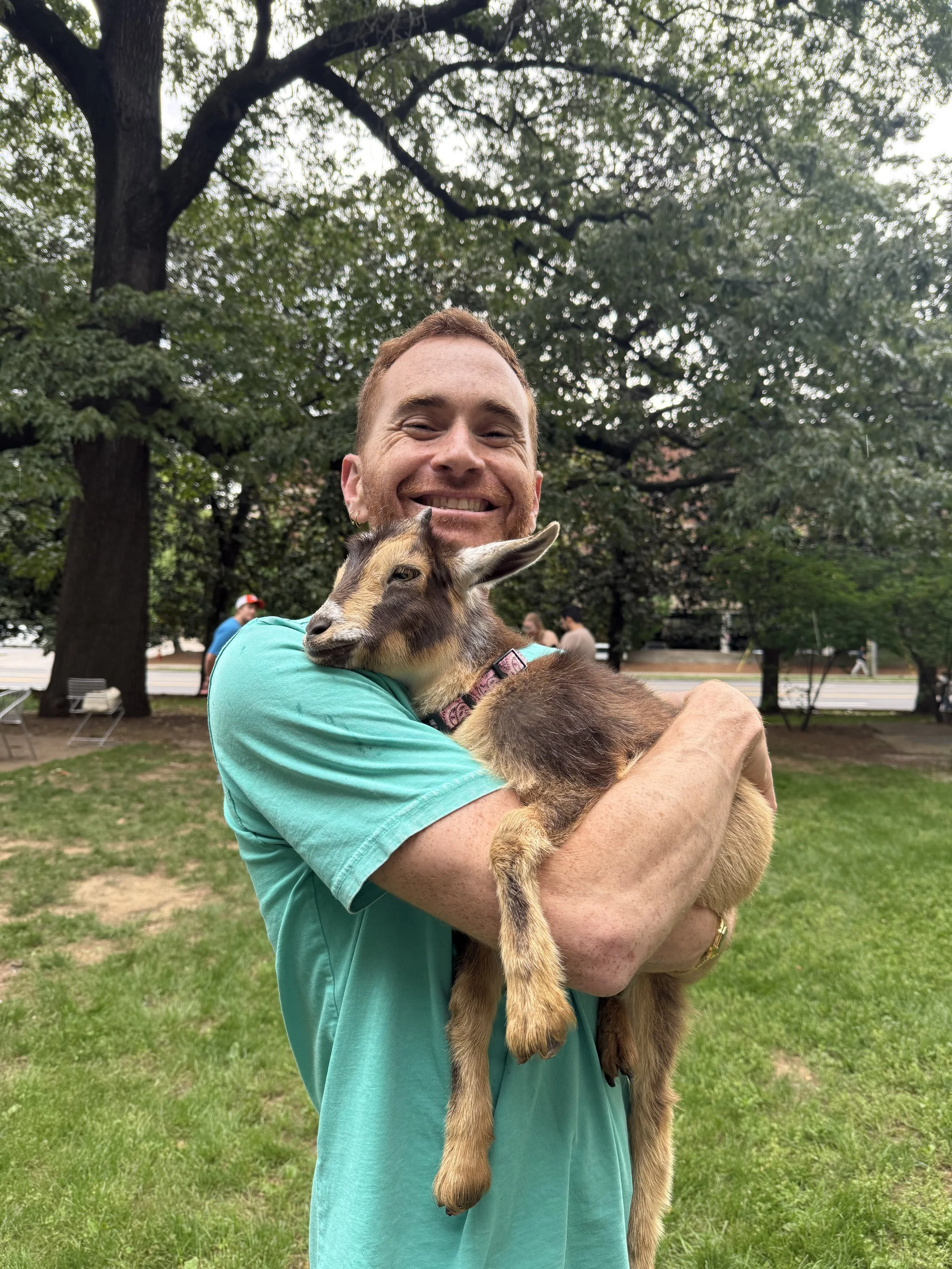 A man with a big smile holding a small goat in his arms outdoors in a park, with large trees in the background.