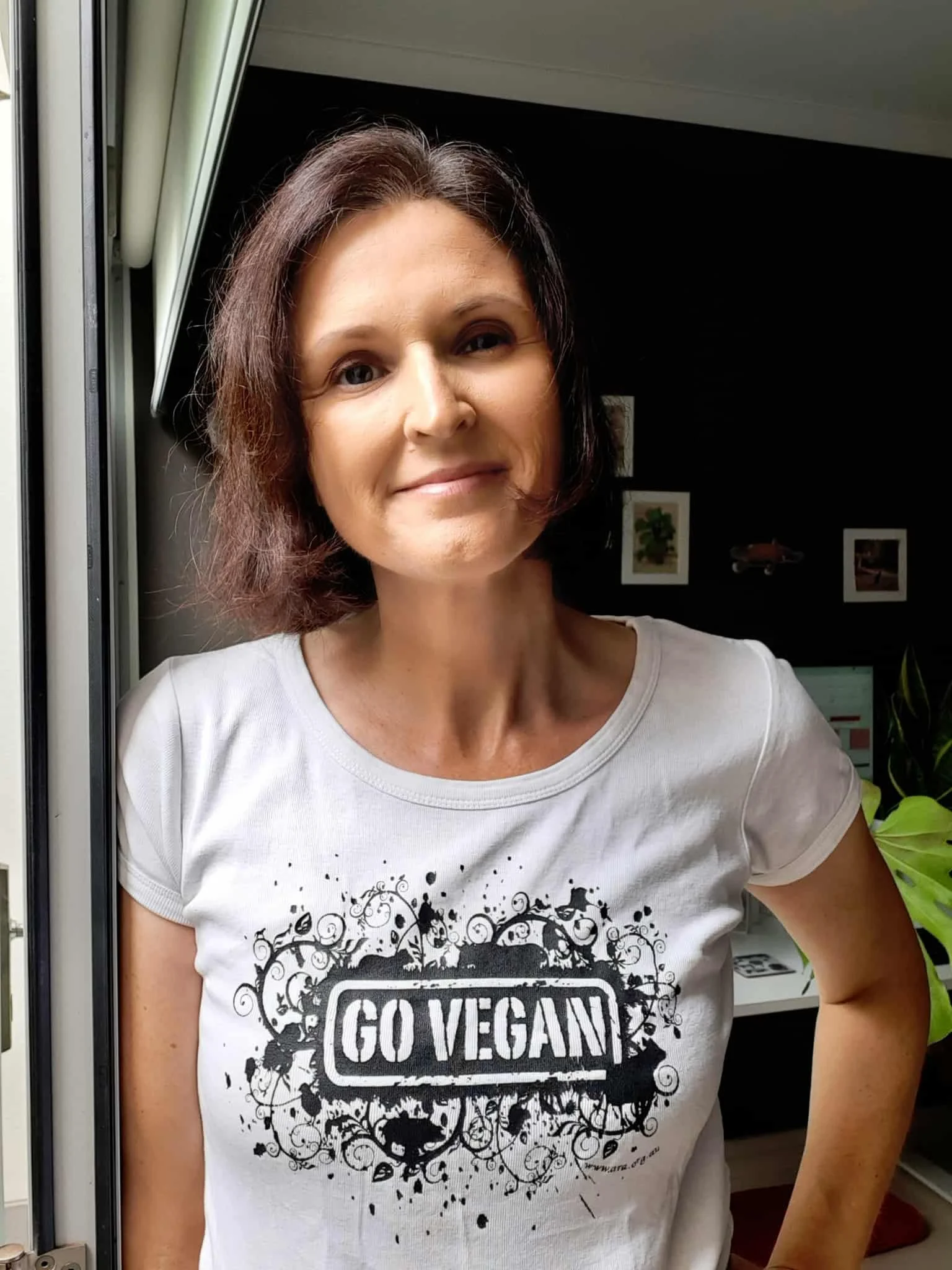 A confident women with a vegan t-shirt saying GO VEGAN in a home background with plants and pictures