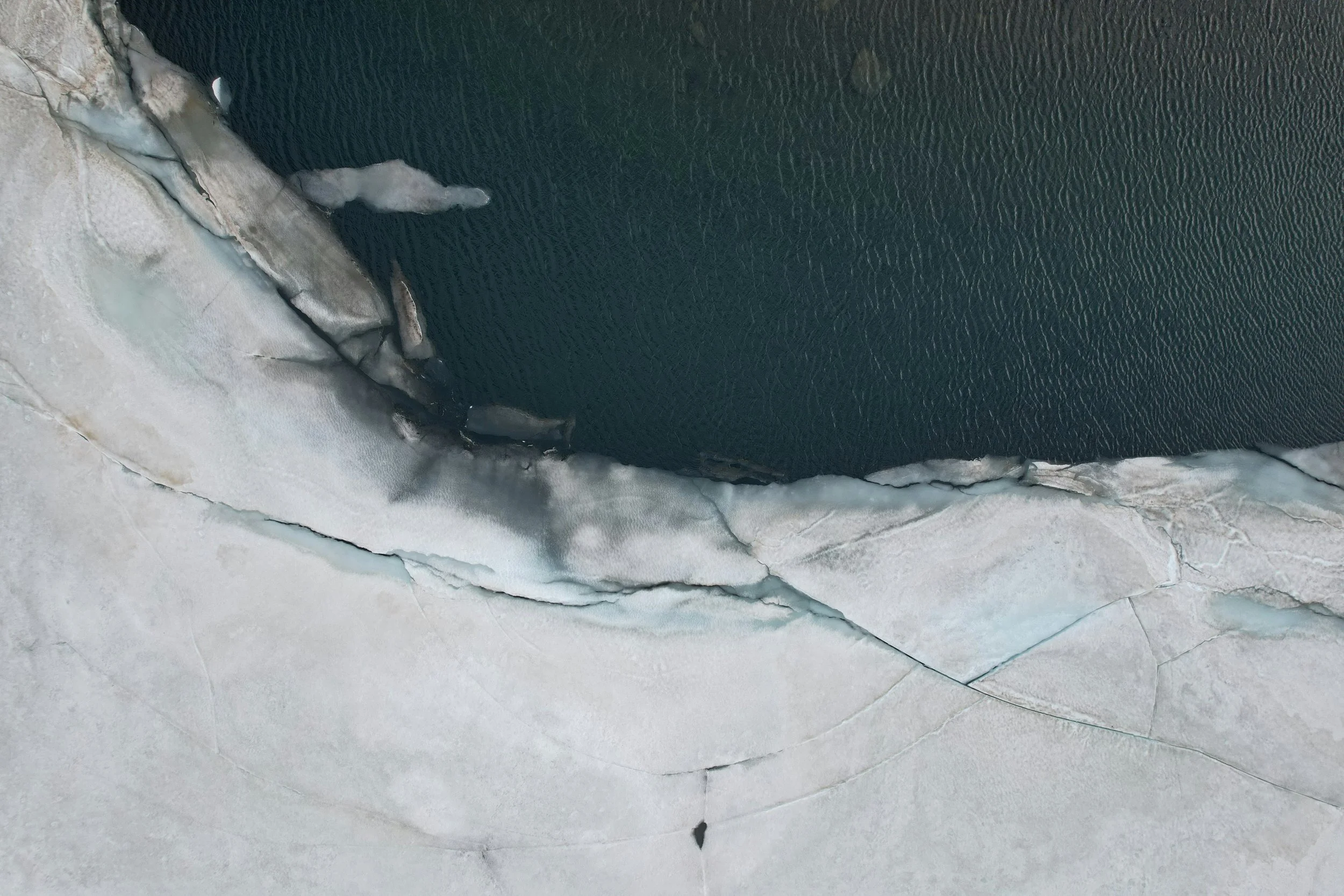 Aerial view of a frozen ice sheet with a dark water or ice-covered surface at the top, and cracked, light-colored ice or snow at the bottom.
