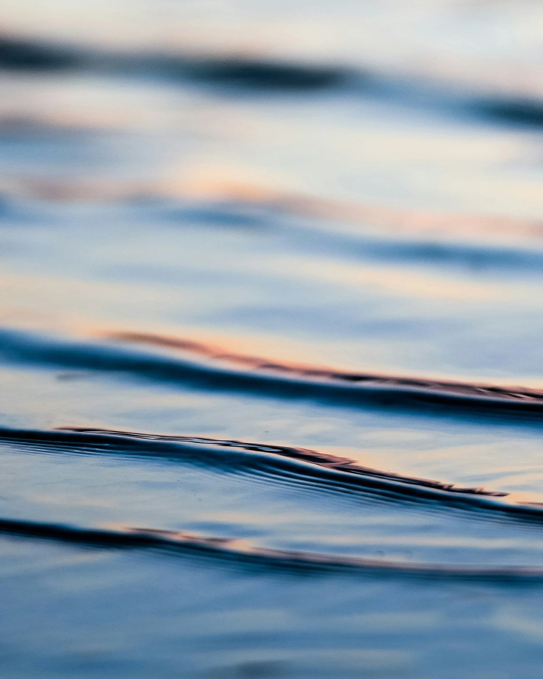 Close-up of calm water with gentle ripples and soft pastel sunset reflections.