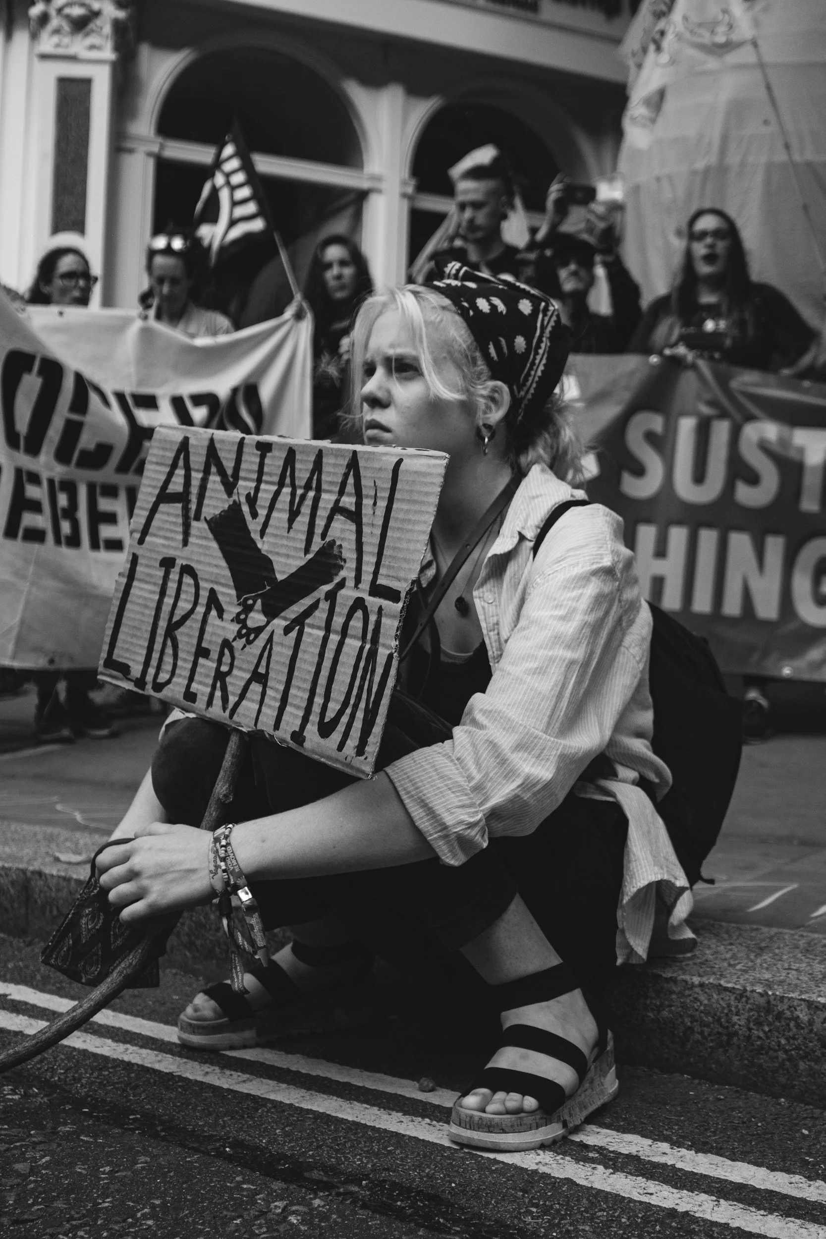 A young woman squatting on the street holding a sign that reads 'Animal Liberation' during a protest, with other protesters and banners in the background.