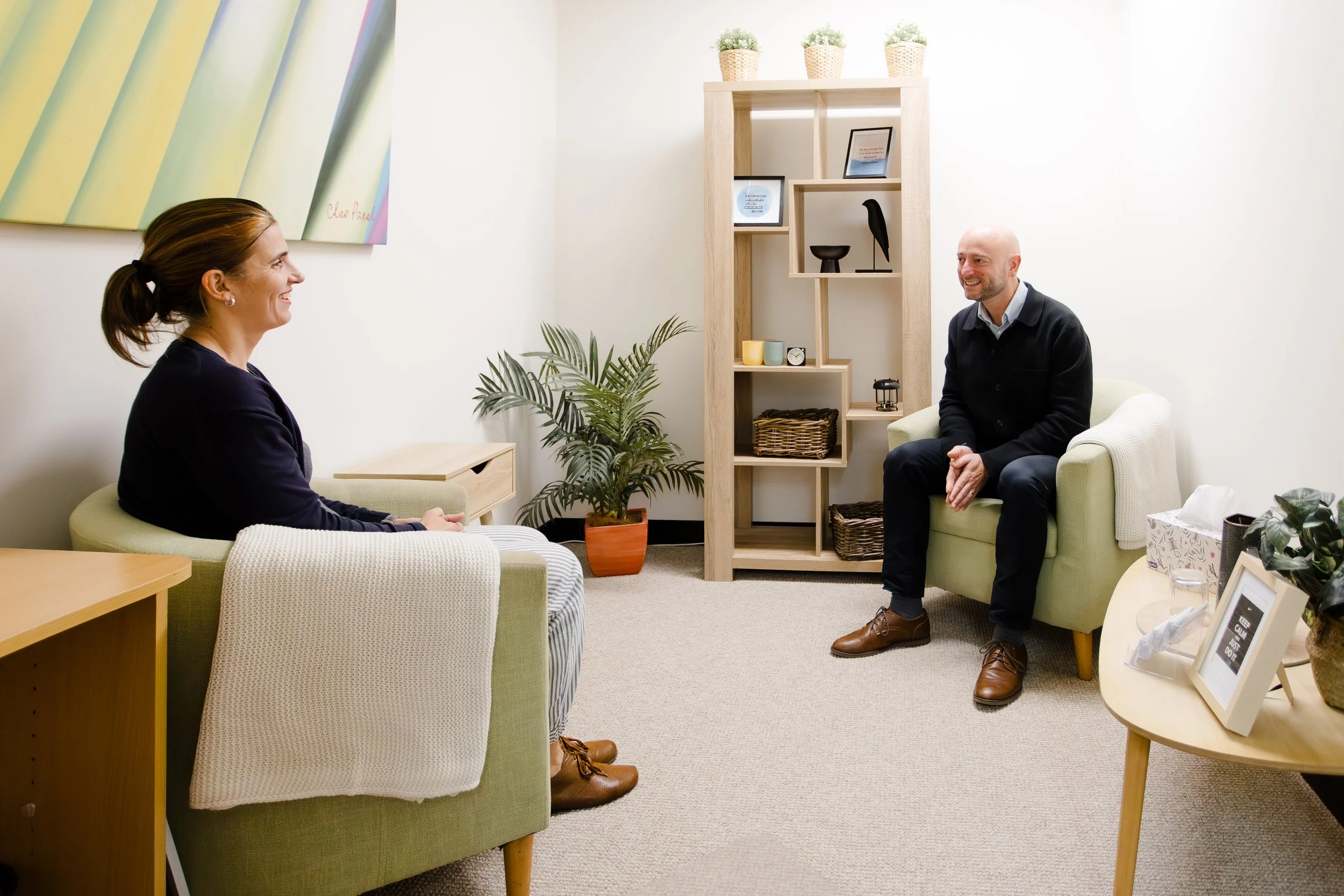 A woman and a man sitting in a therapy or counseling office, facing each other, smiling and engaging in conversation. The room has light-colored walls, a plant, and a wooden shelf with decorative items, picture frames, and baskets.