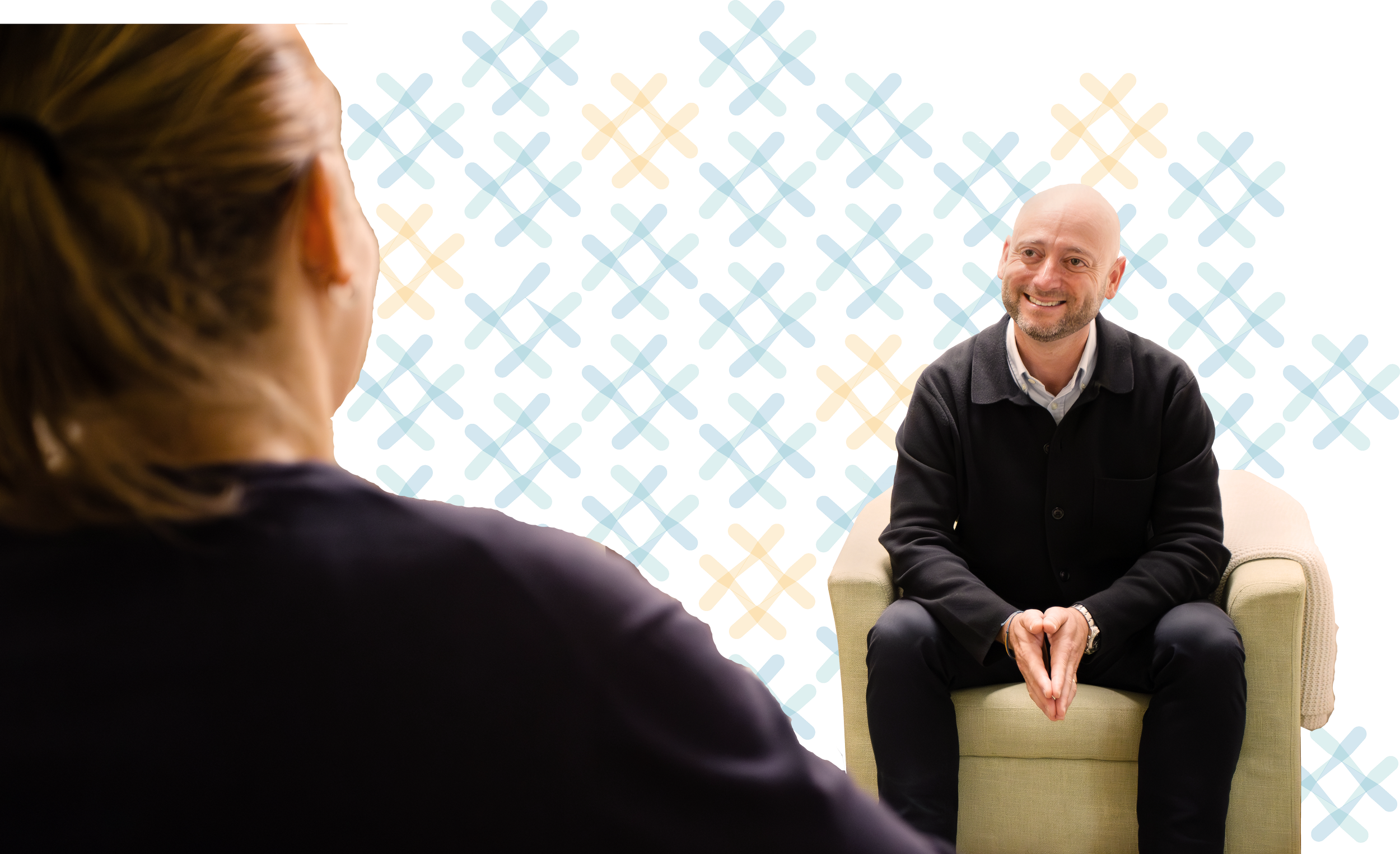 A man with a bald head and beard smiling, sitting on a light-colored armchair, talking to a woman with brown hair, in front of a colorful patterned background.