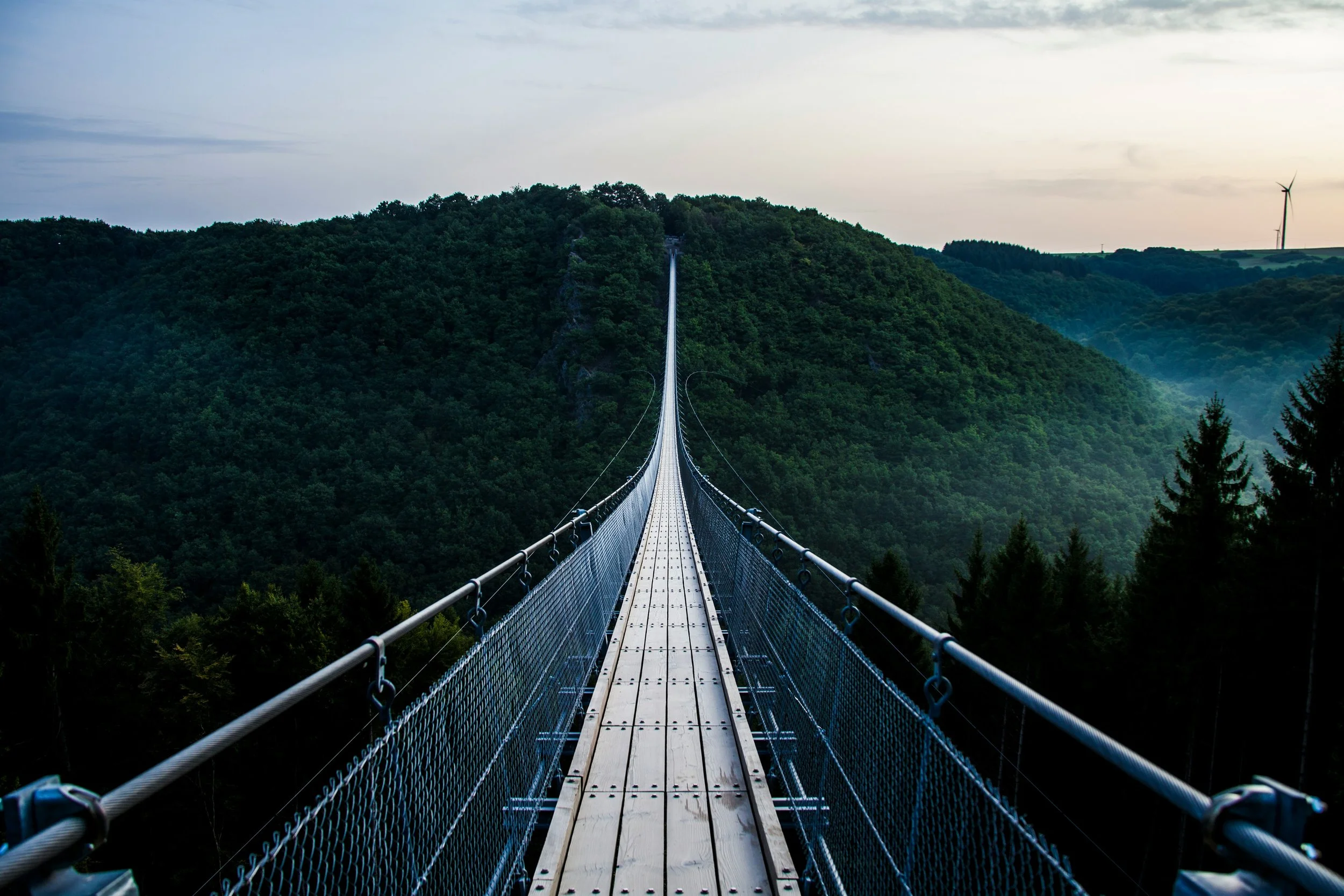 A long suspension bridge extending over a deep green forested valley with hills in the distance, under a cloudy sky.