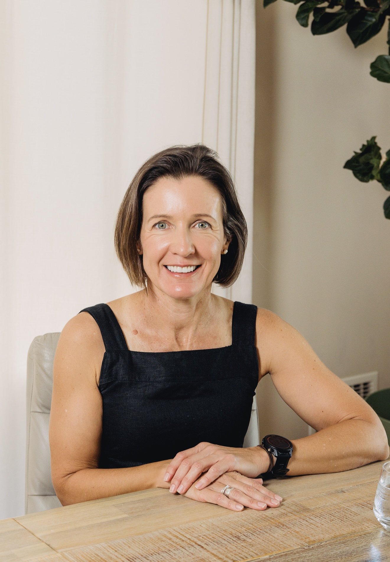 A woman with short brown hair smiling, wearing a black sleeveless top and a wristwatch, sitting at a wooden table with hands folded, in a room with beige walls and a large leafy plant in the background.