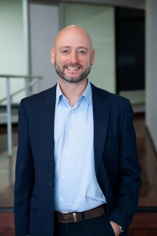 Bald man with a beard in a navy suit and light blue shirt smiling in an indoor office setting.