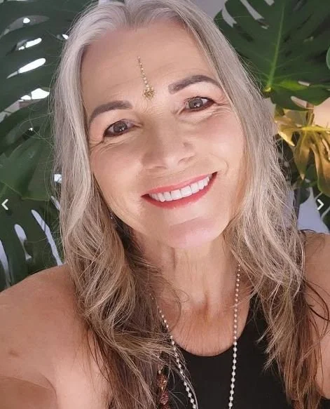 A smiling woman with gray hair and a jeweled forehead adornment, wearing a black top and a pearl necklace, standing in front of large green tropical leaves.