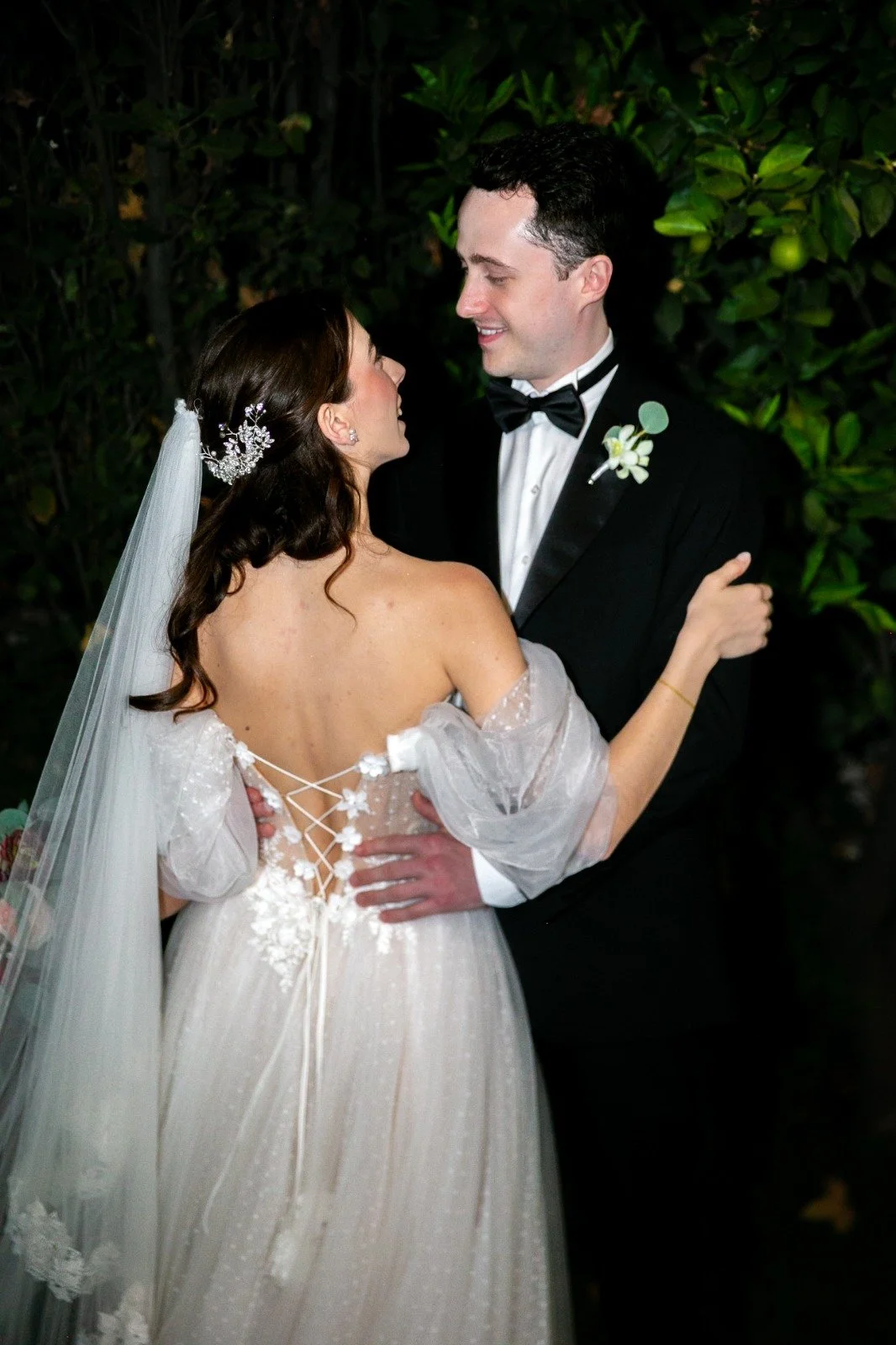 Una pareja vestida con ropa de boda, la mujer con vestido blanco y el hombre con traje negro, compartiendo un momento romántico en un entorno natural con vegetación de fondo.