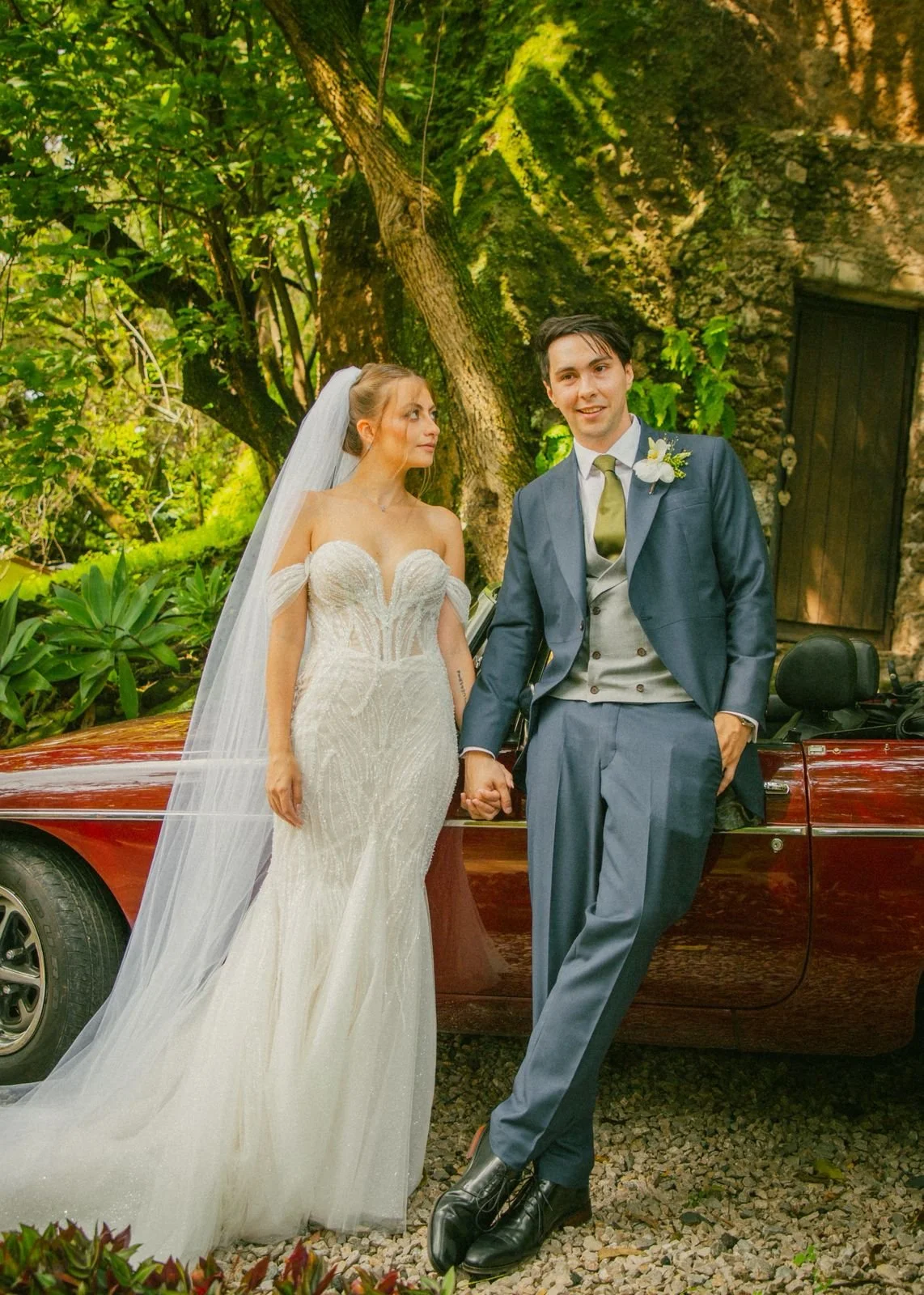 Una pareja de recién casados, la mujer con vestido de novia y el hombre con traje, están tomados de la mano frente a un coche de color burdeos, rodeados de naturaleza y un árbol grande en un entorno natural.