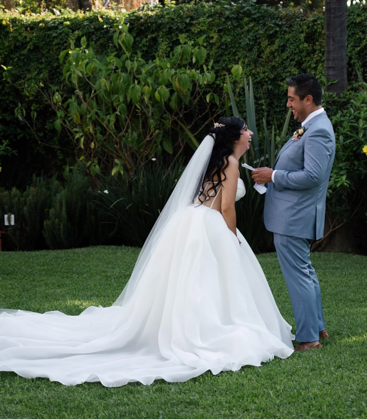 Pareja de recién casados en jardín, la novia con vestido largo y velo, el novio con traje azul, ambos riendo y mirándose con alegría.
