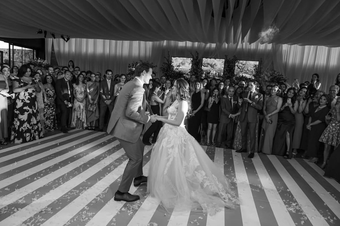 Pareja de novios bailando en el centro de una boda, frente a un grupo de invitados que los observa y toma fotos, en un espacio decorado con cortinas y plantas.