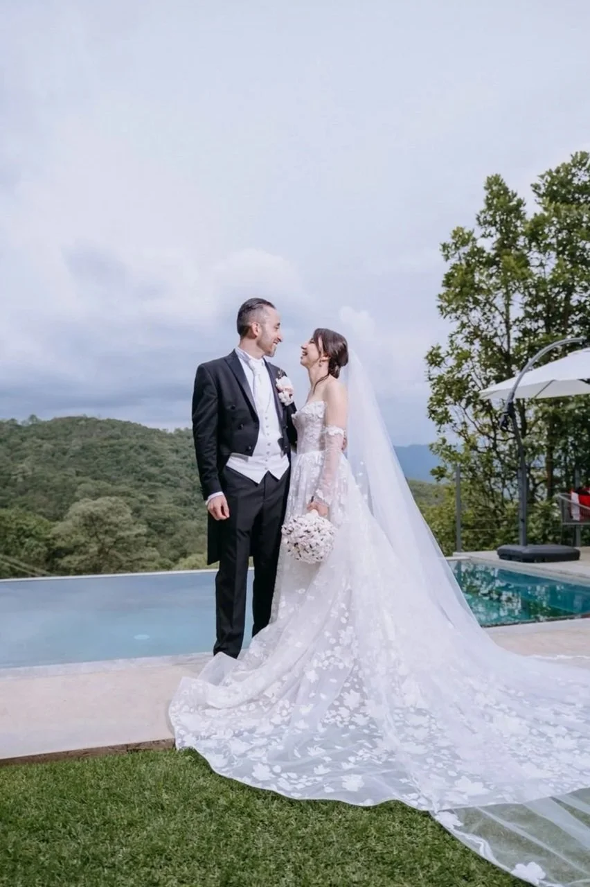 Pareja de novios en boda frente a una piscina, con fondo de montañas y árboles, ambos sonriendo y mirando fijamente.