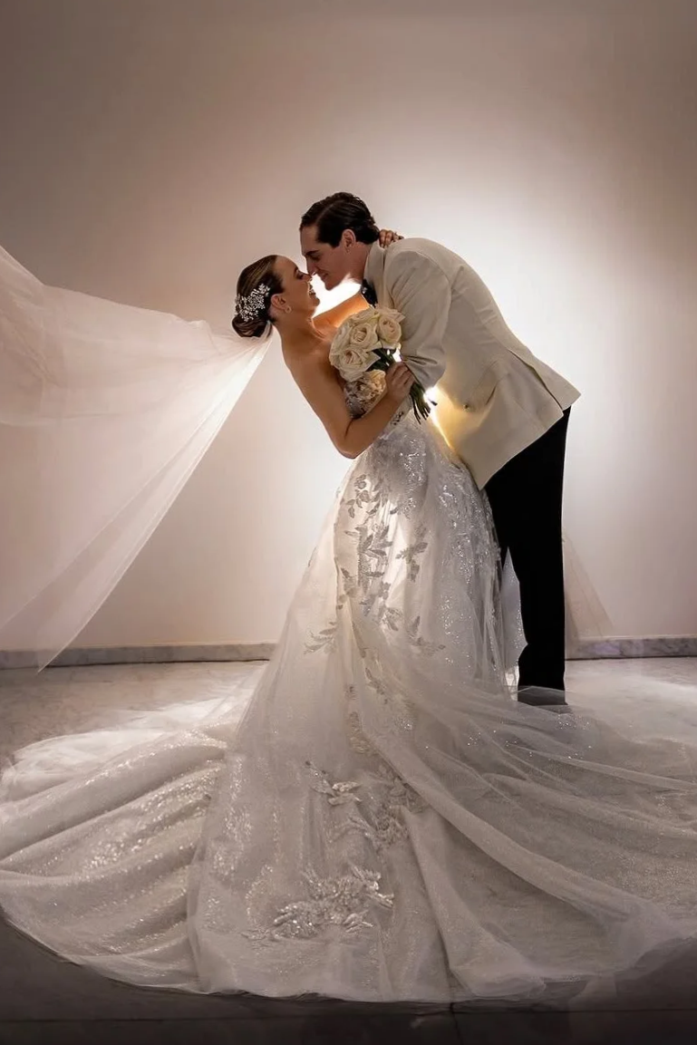 Novios abrazados, en una foto de boda, ambos con sonrisas, la novia con vestido largo y velo, el novio con saco blanco y pantalones oscuros, en un fondo neutro con iluminación suave.