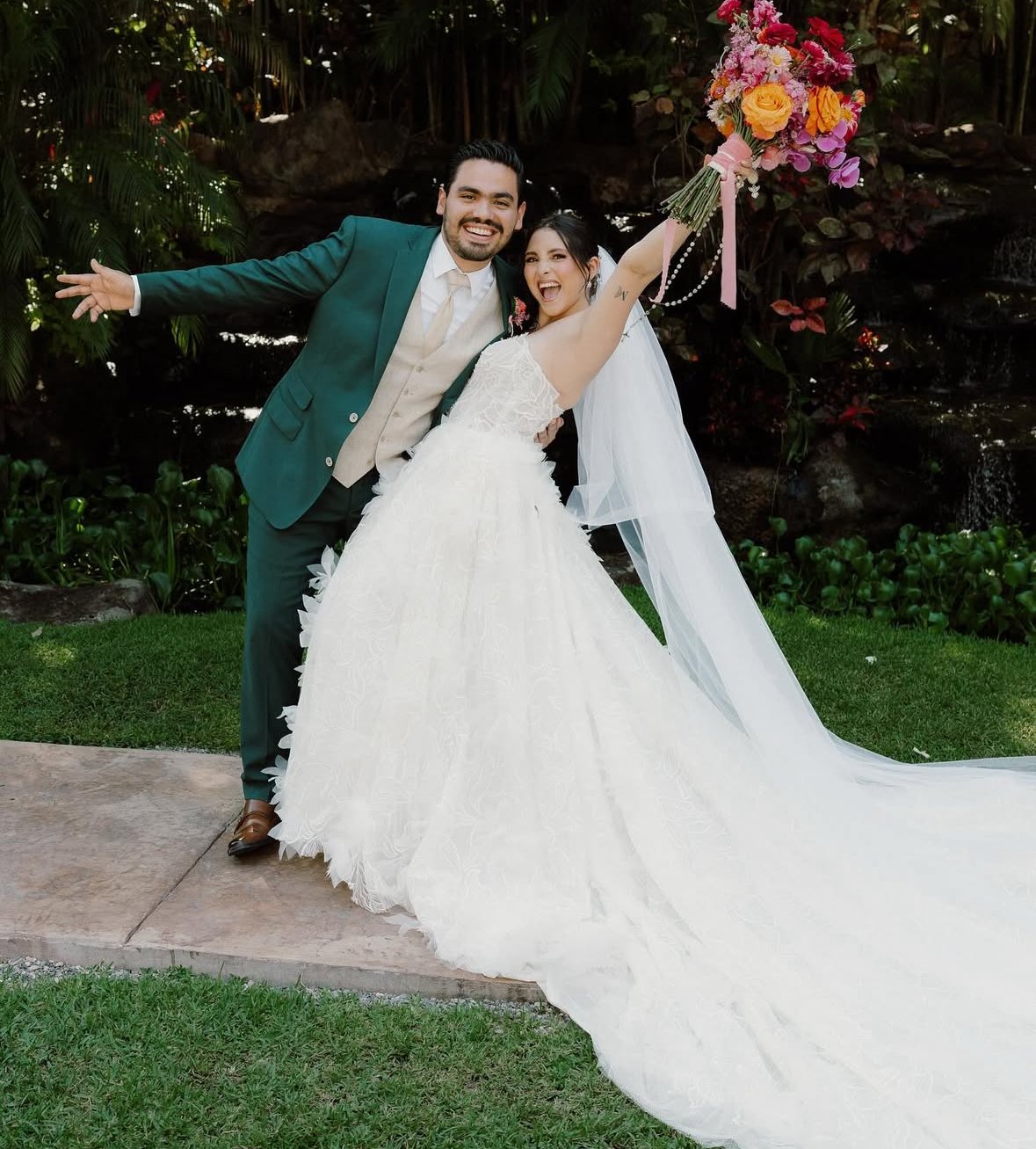 Pareja de recién casados, la novia con vestido blanco y velo, sosteniendo un ramo de flores, el novio con traje verde, en un jardín con plantas y una pequeña cascada en el fondo.