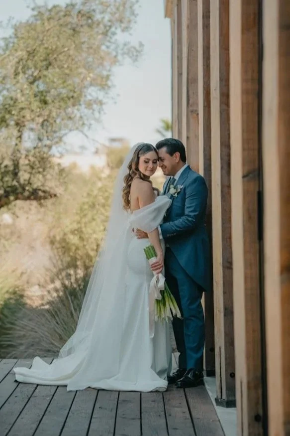 Pareja de recién casados en un entorno exterior, con la novia sosteniendo un ramo de flores y vestida con vestido de novia y velo, mientras el novio se viste con traje azul, ambos sonriendo y en actitud romántica.