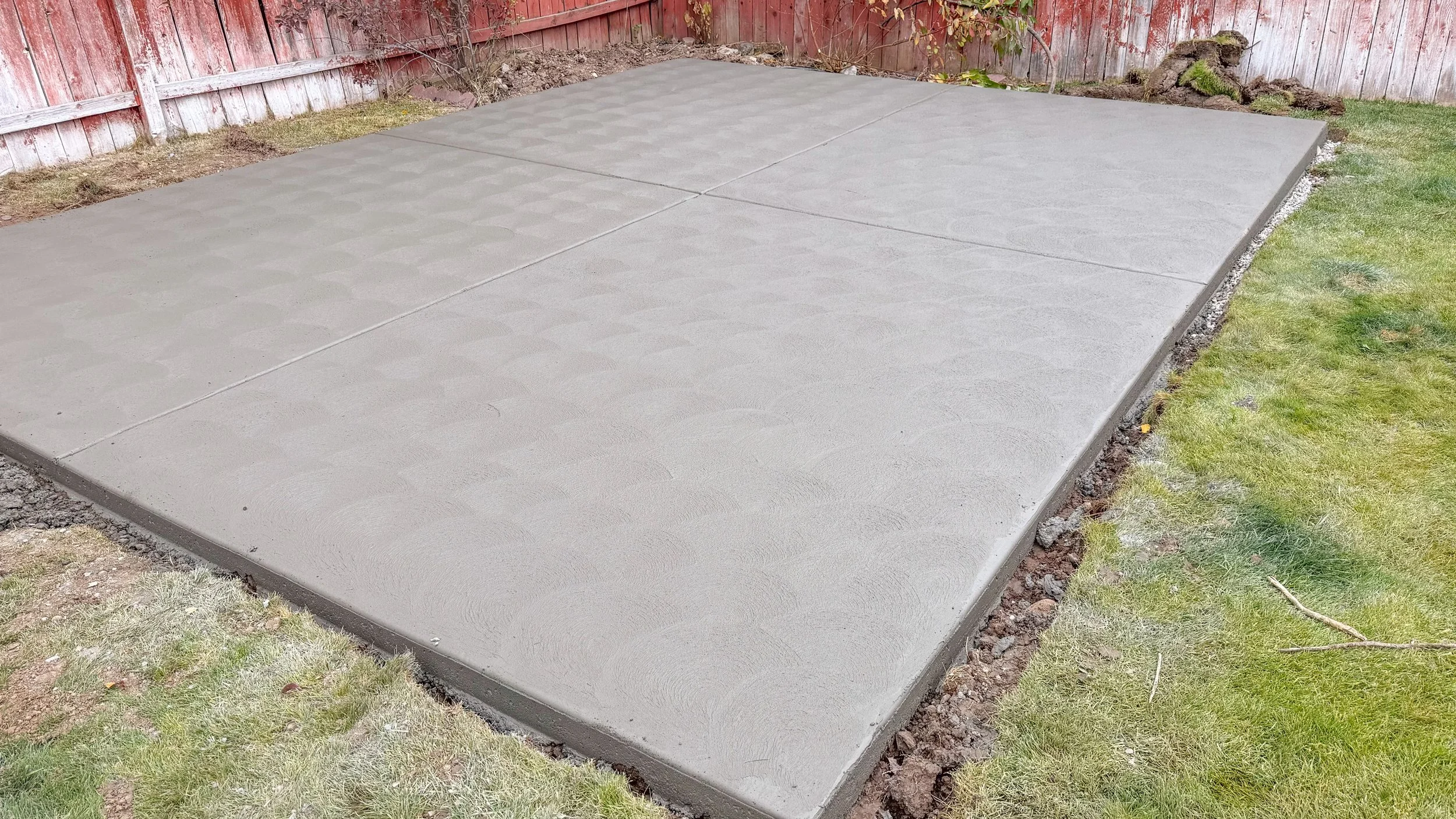 Freshly poured concrete slab in a backyard surrounded by grass, a wooden fence, and garden soil.