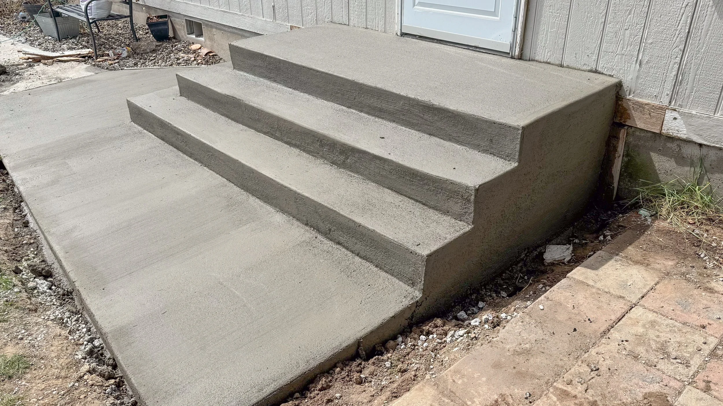 Concrete front steps leading up to a door, with three stairs and a small landing, next to a house with siding, and some grass and dirt around the edges.