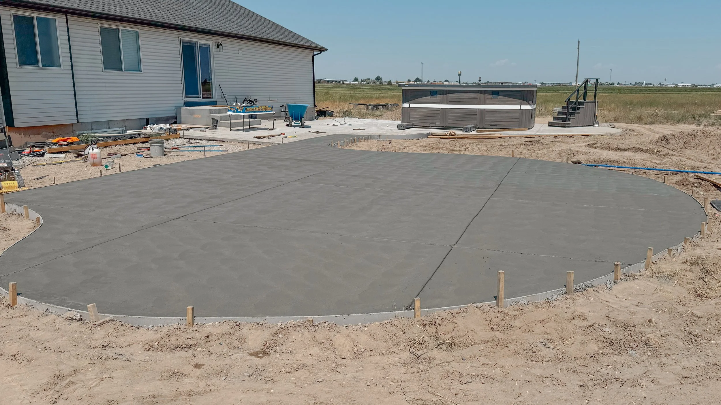 Freshly poured concrete slab with curved edges in a backyard under construction, with house and construction tools in the background.