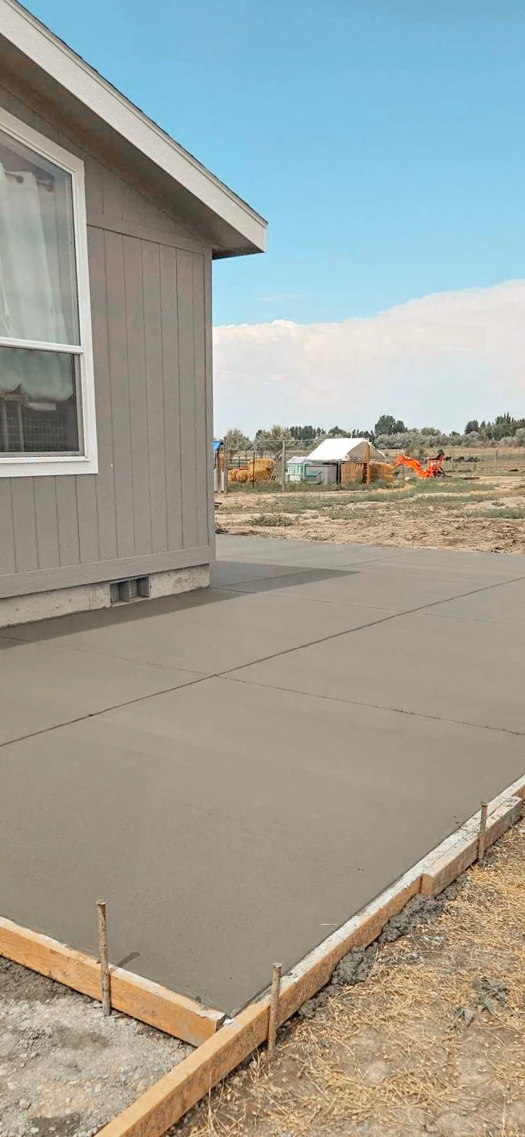 Freshly poured concrete slab in a backyard, bordered by wooden forms, with part of a house and construction site visible in the background under a clear sky.