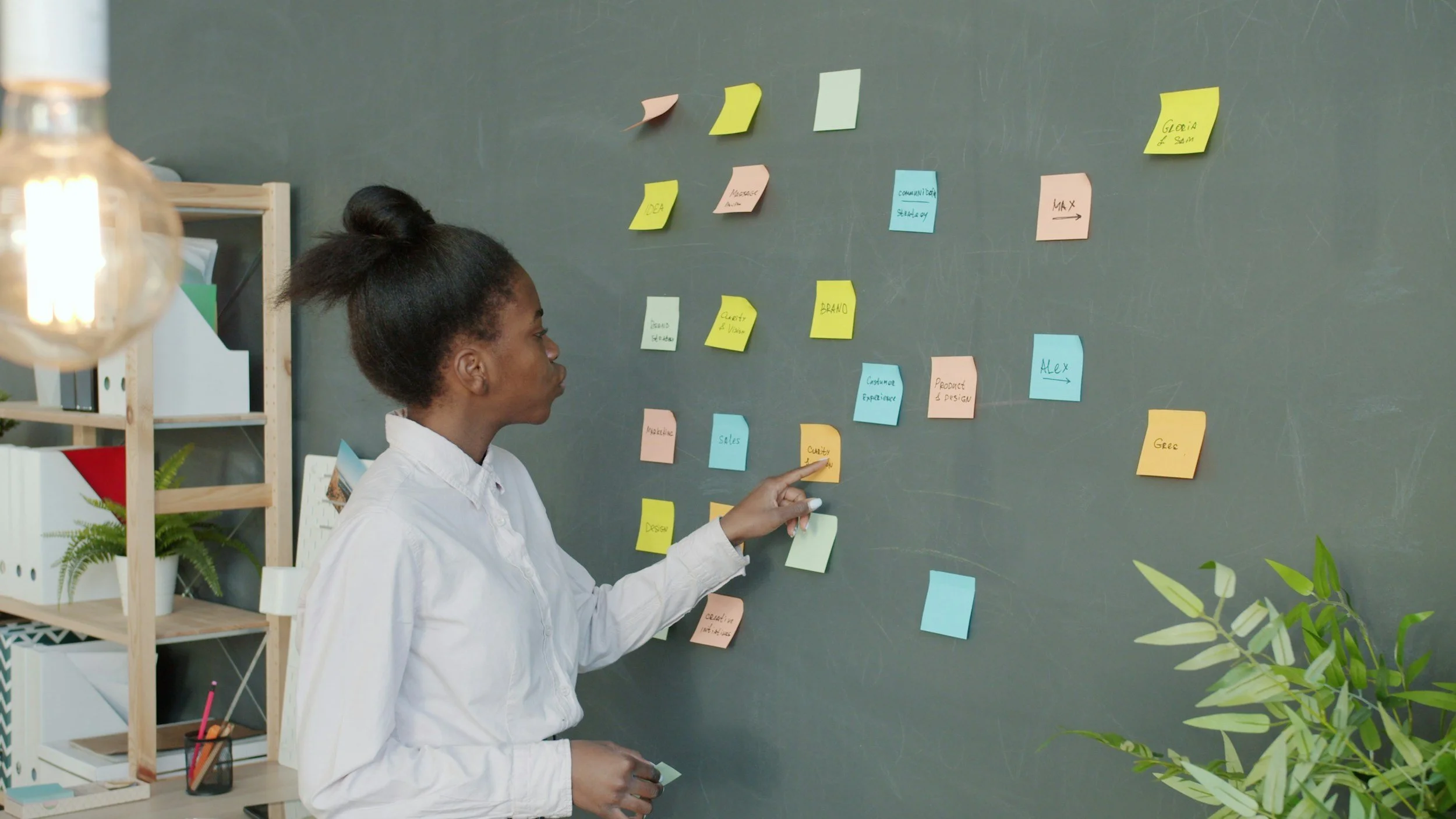 Woman in white shirt placing yellow sticky note on a blackboard covered with colorful sticky notes, in a workspace with shelves and plants.