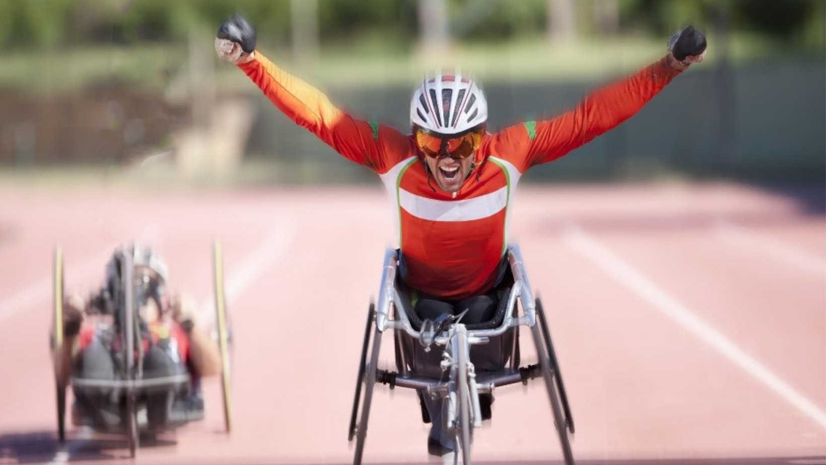 A male athlete in a racing wheelchair celebrating with arms raised on a track, wearing a red and white sports outfit, helmet, and sunglasses, with another racing wheelchair in the background.