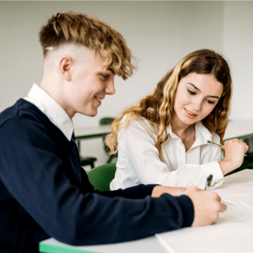 Two students sitting at a desk in a classroom, writing in notebooks and engaging with each other.