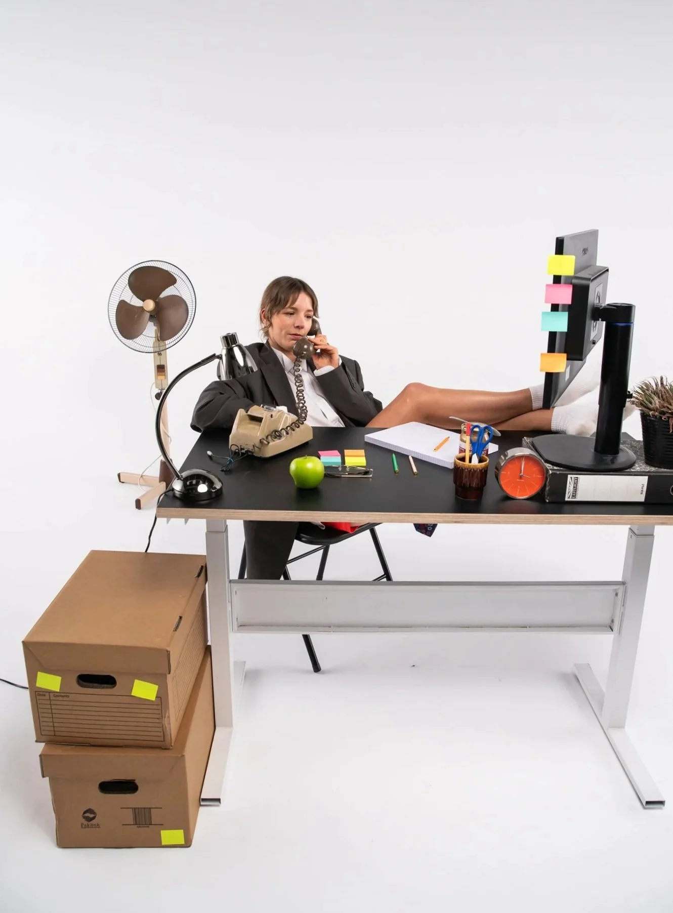A woman in professional attire sitting at a cluttered desk with her feet up on the computer monitor, talking on the phone, surrounded by office supplies, a small fan, and cardboard boxes.