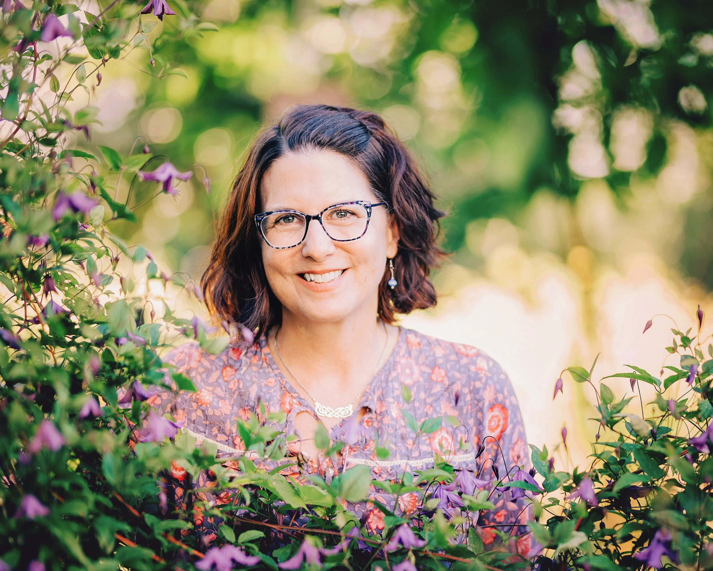 A woman with shoulder-length dark hair, wearing glasses and earrings, smiling outdoors among green foliage and purple flowers, with sunlight filtering through the background.