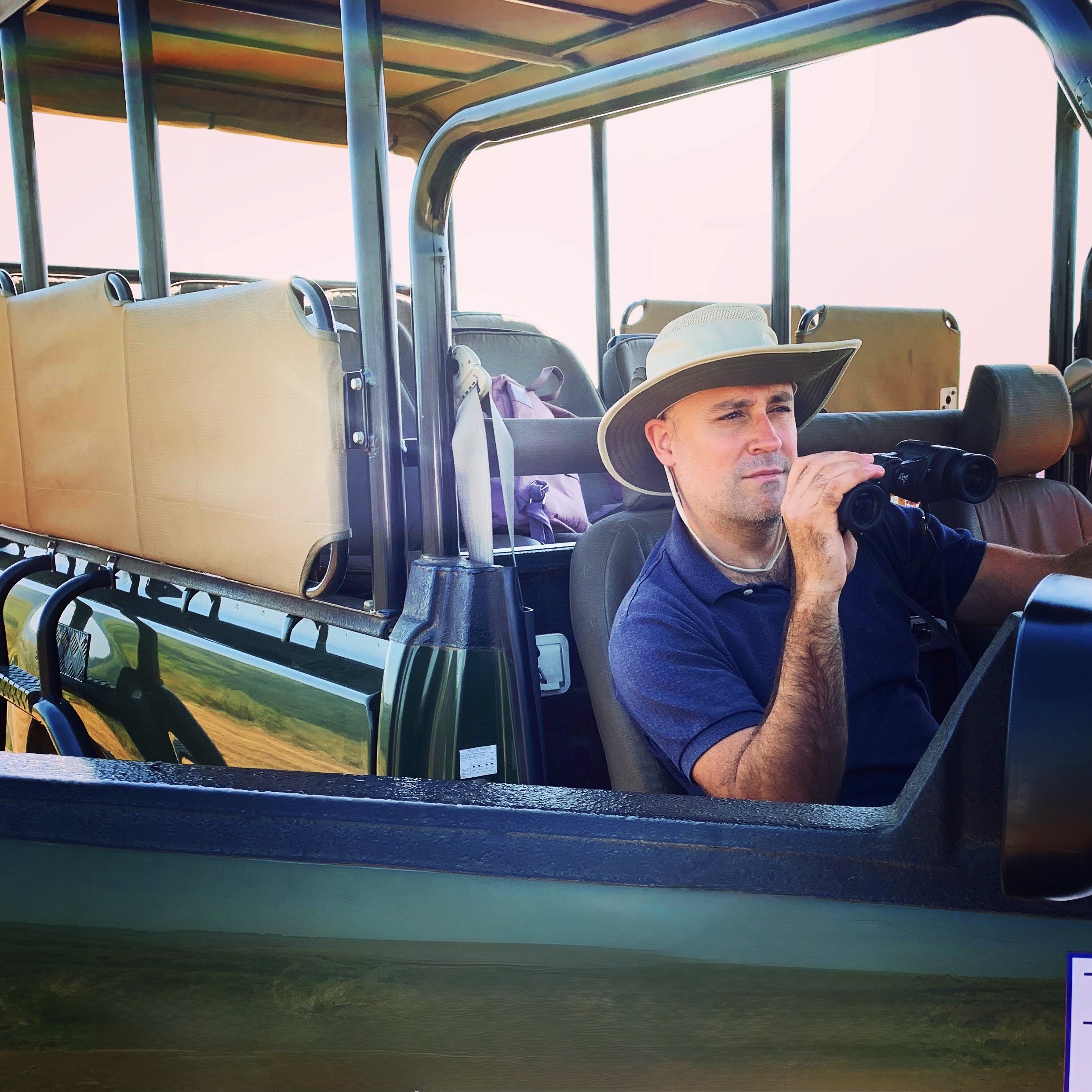 A man wearing a safari hat and a navy polo shirt sits in a safari vehicle, holding binoculars and looking intently into the distance on a safari. The vehicle has a roof and open sides, with seats and some backpacks visible in the background.