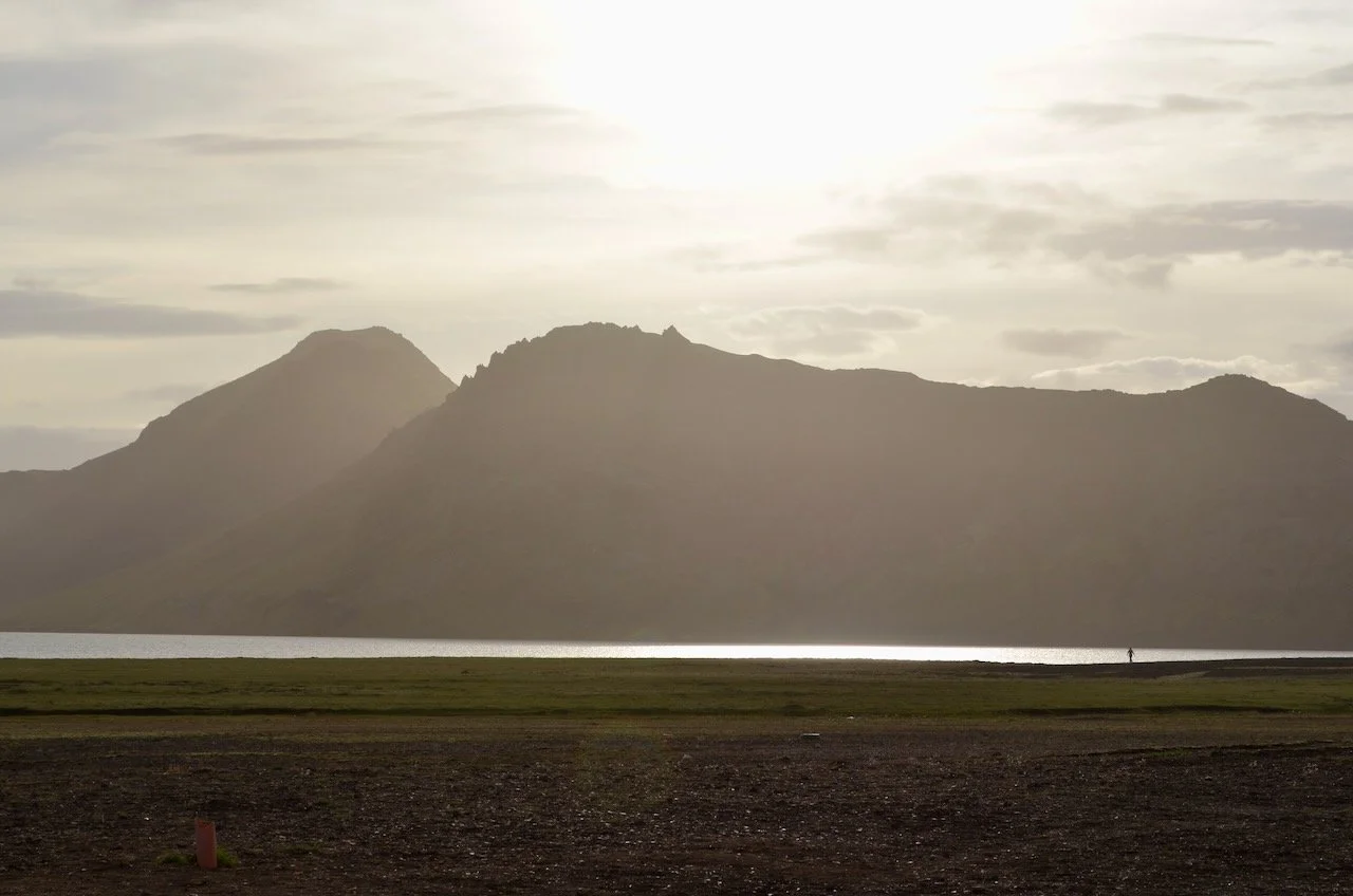 Scenic landscape with mountains in the background, a body of water, and a person walking along the shoreline during sunset.