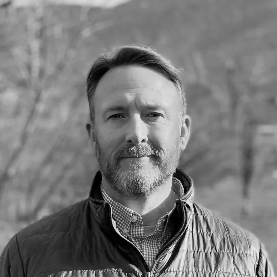 Black and white portrait of a man with a beard, short hair, wearing a quilted jacket and a collared shirt, outdoors with blurred trees in background.