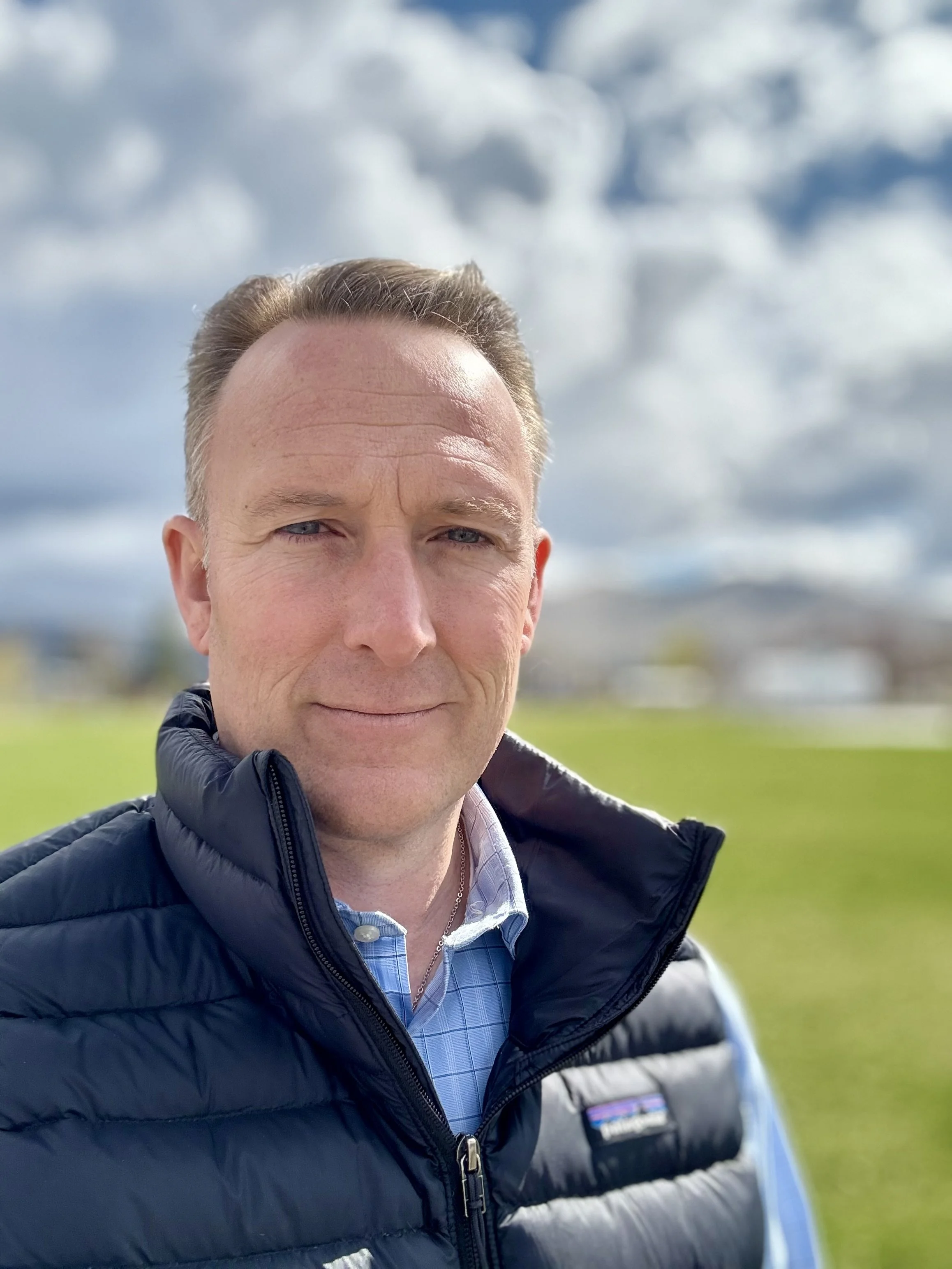 A man outdoors in a grassy area with cloudy sky in the background. He is wearing a black puffer vest over a blue button-up shirt.
