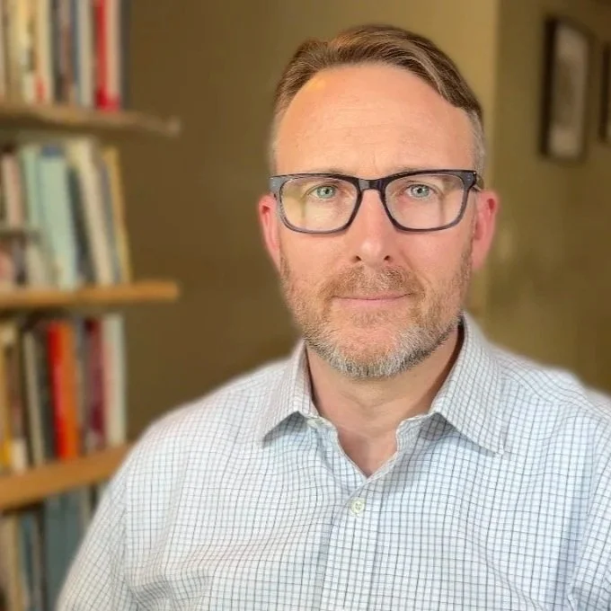 A man with glasses and a beard, wearing a checkered shirt, standing in a room with bookshelves in the background.